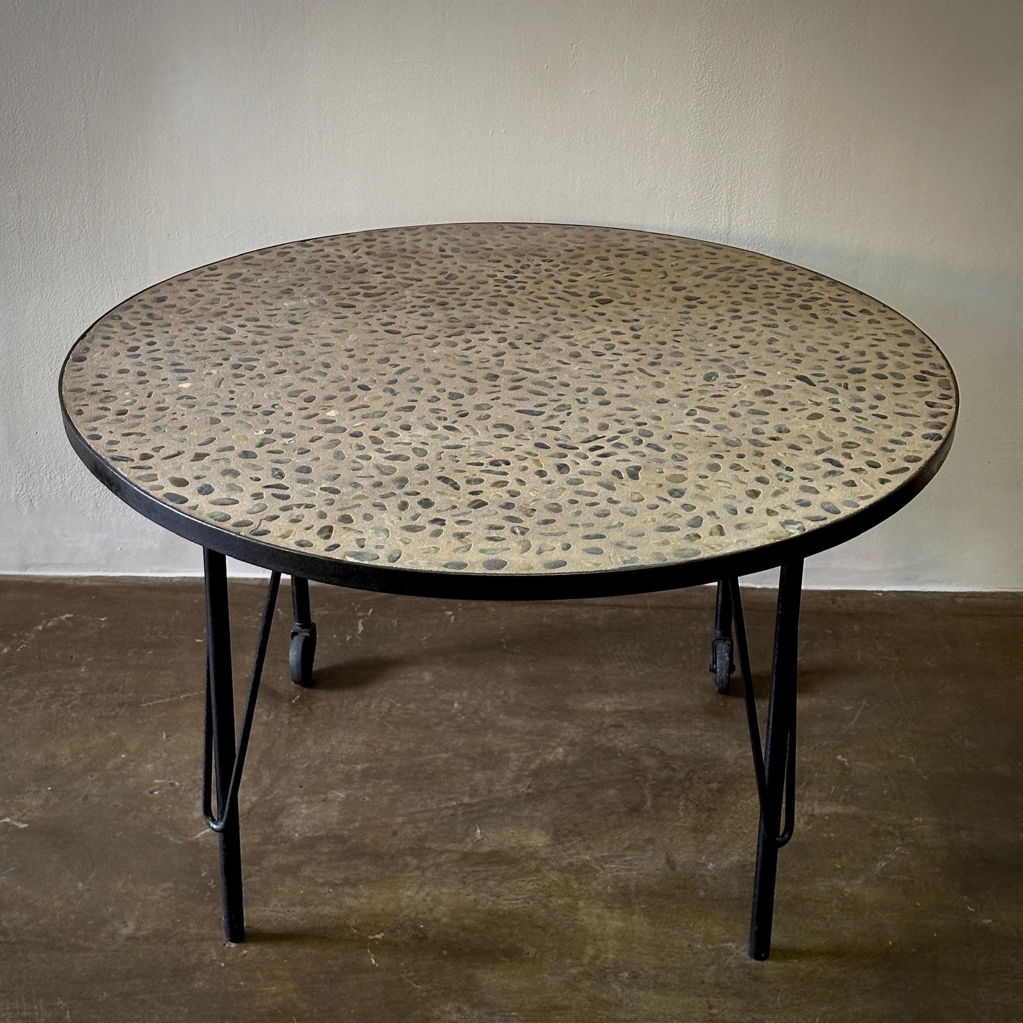 Cette table anglaise marie magnifiquement les éléments industriels et naturels, avec une base ronde en fer et un plateau en béton orné d'une incrustation de pierre de rivière naturelle. Le motif organique des pierres par rapport au cadre austère et