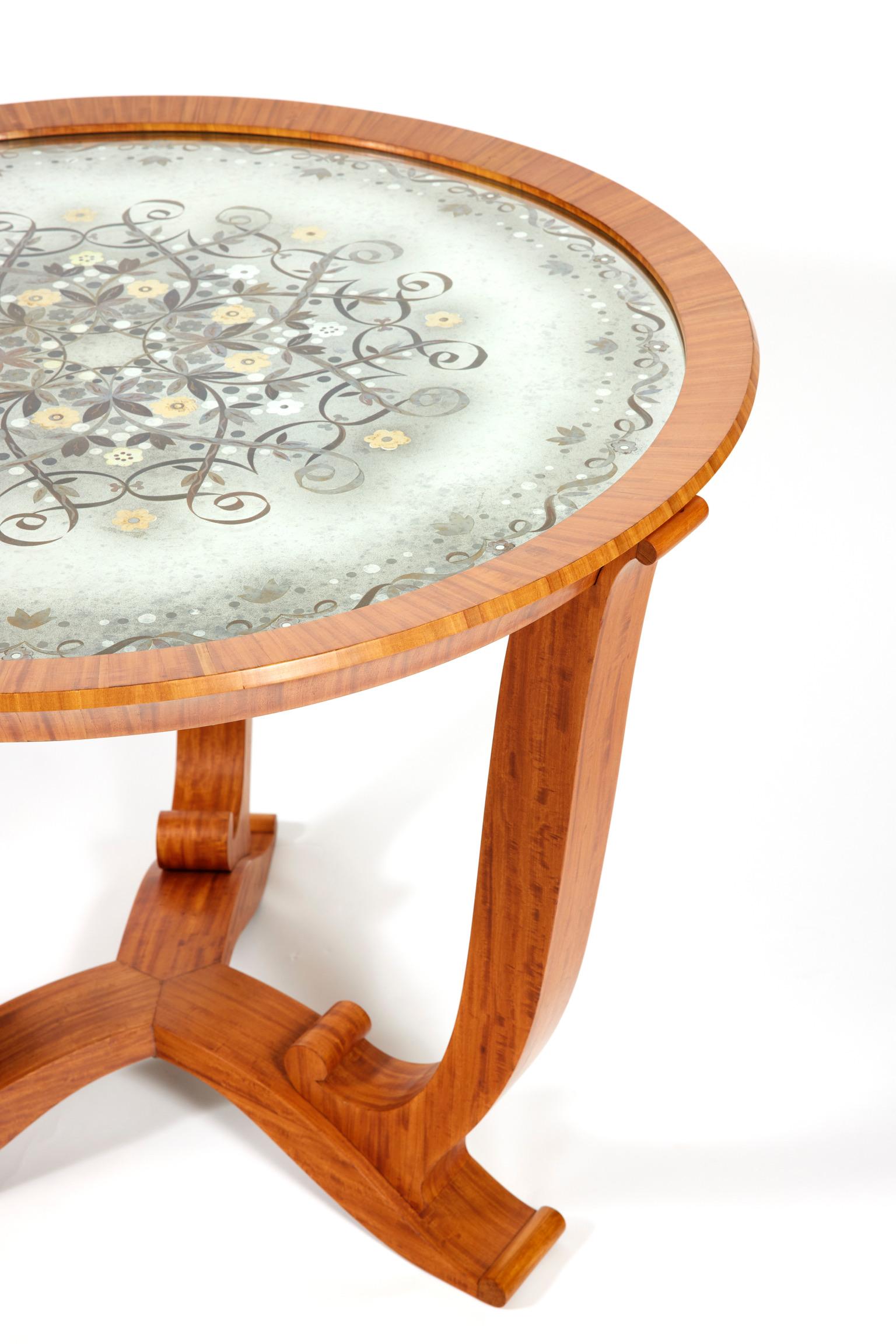 Lemon tree veneer pedestal table with a tripod base and console-shaped feet.
The circular églomisé glass top is decorated with scrolls and stylized flowers in golden leaves.
Signed on an ivory plaque “J. Leleu”.