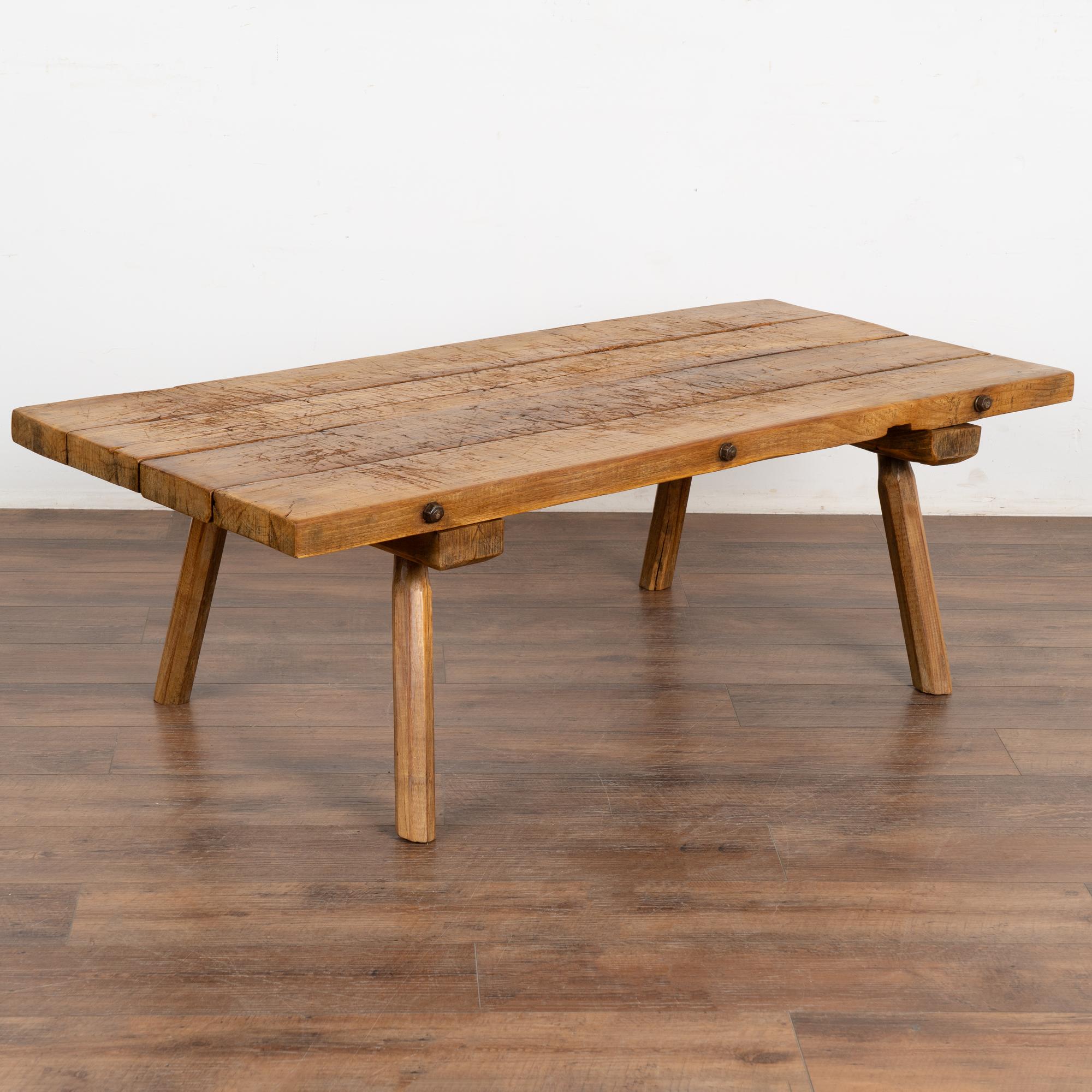This rustic slab wood coffee table with peg legs is loaded with vintage character due to the heavily distressed wood of the top and visible iron bolts along the edge.
The thick top made of four heavy planks is covered in scrapes, gouges, cracks,