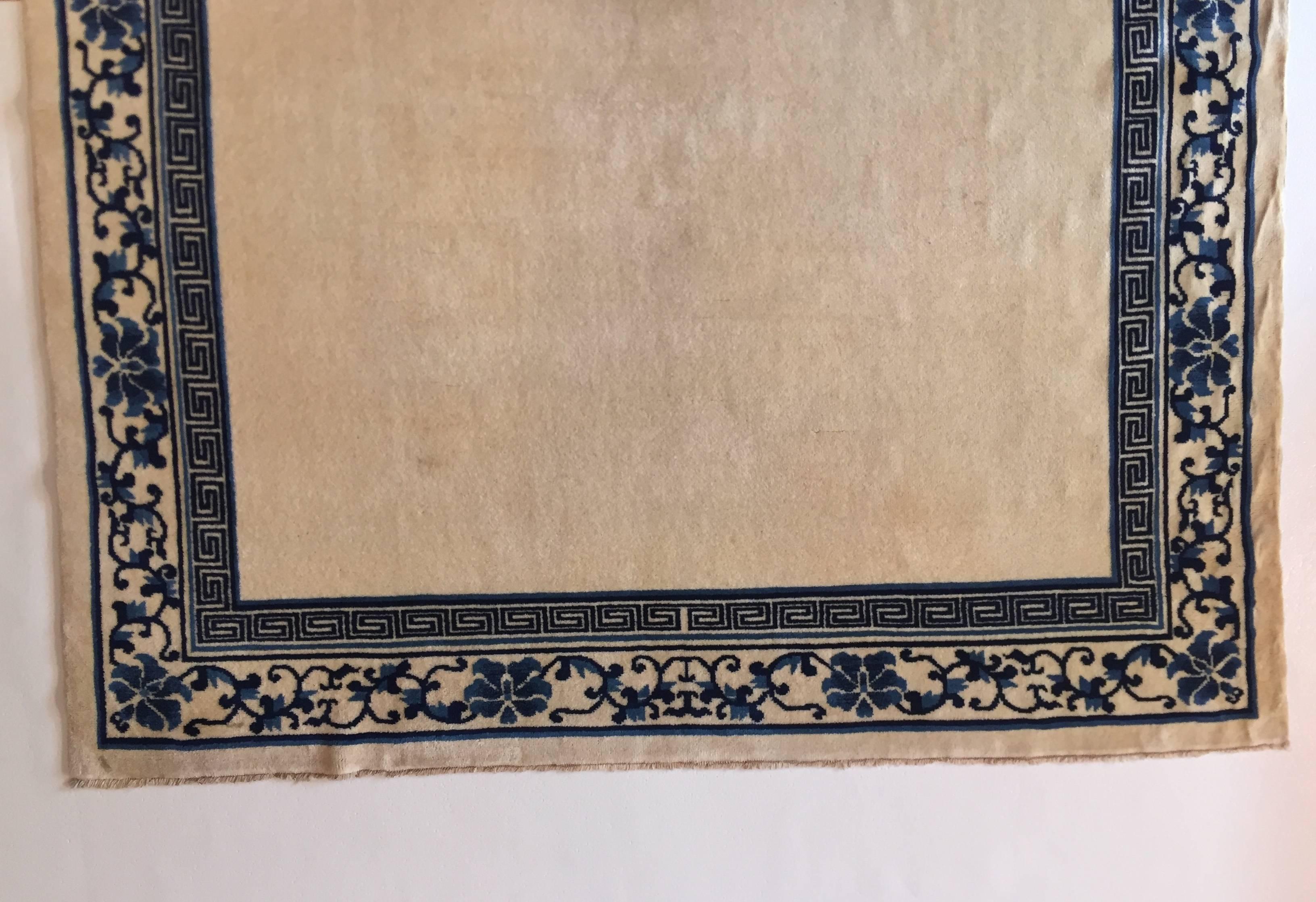 Tapis de Pékin de couleur crème. Bordures dans d'autres tons de bleu décorées d'une rangée de fleurs de lotus soulignées par un grec. Un produit typique de l'artisanat chinois inspiré de la porcelaine blanche et bleue de la dynastie Ming.