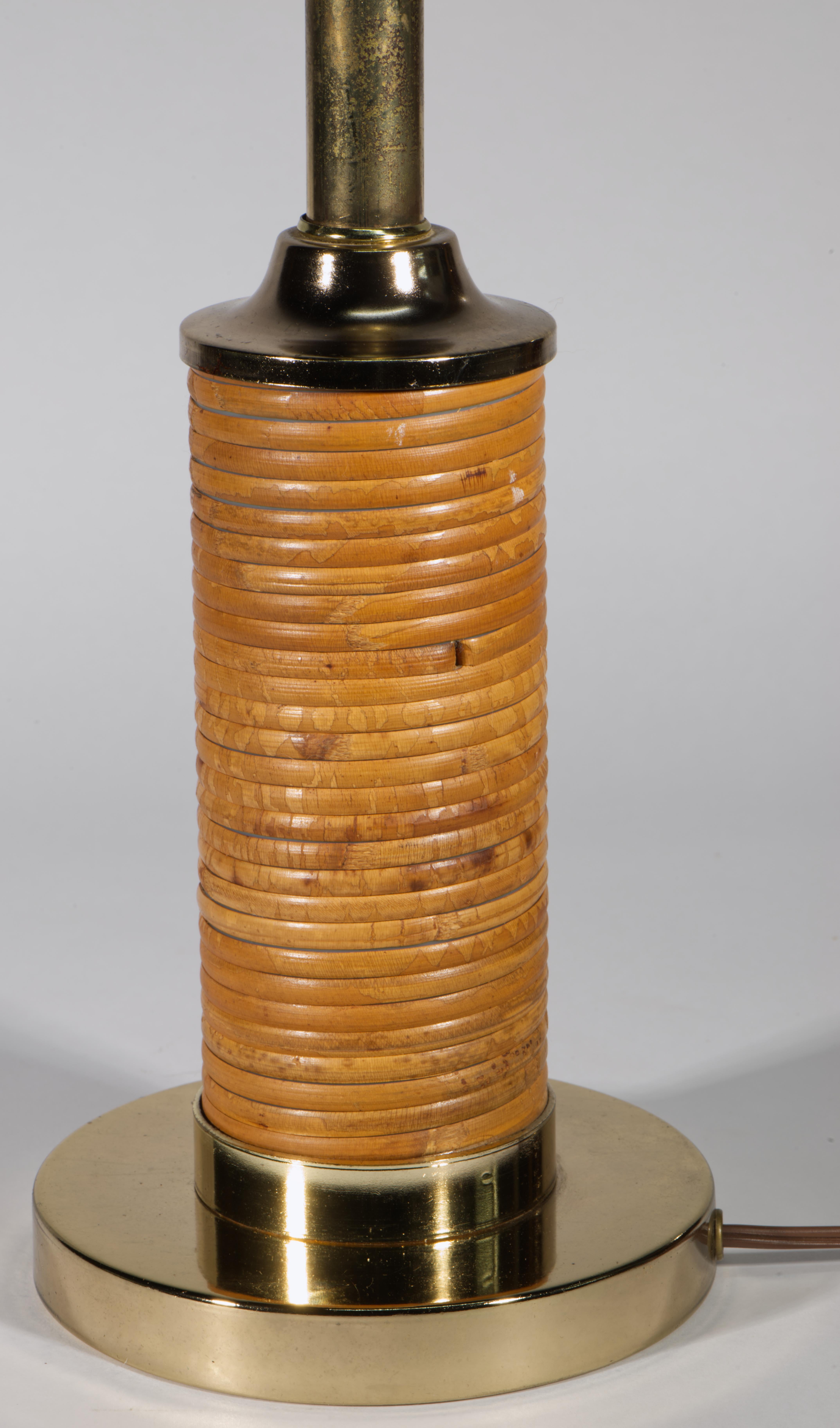 La lámpara de sobremesa vintage de ratán y lat�ón tiene forma de balaustre aerodinámico con cuerpo cilíndrico envuelto en ratán de caña lápiz y montado sobre una base redonda de latón. La parte superior redonda escalonada añade altura y guía la