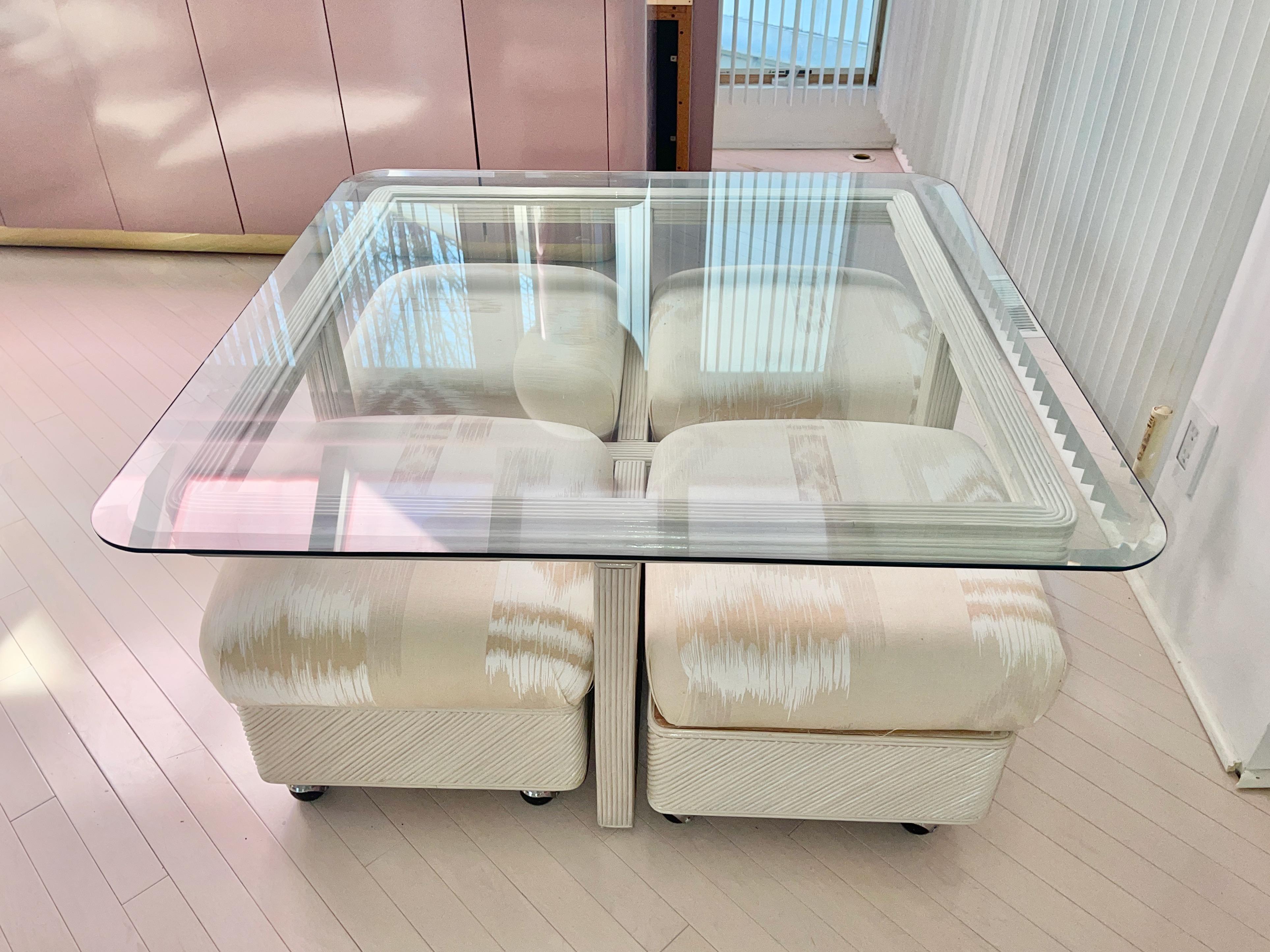 Tavolino da caffè quadrato con piano in vetro in giunco bianco con quattro sgabelli quadrati su rotelle infilati sotto. Anche gli sgabelli sono in giunco bianco con cuscini imbottiti. Il piano in vetro ha uno spessore di 1/2