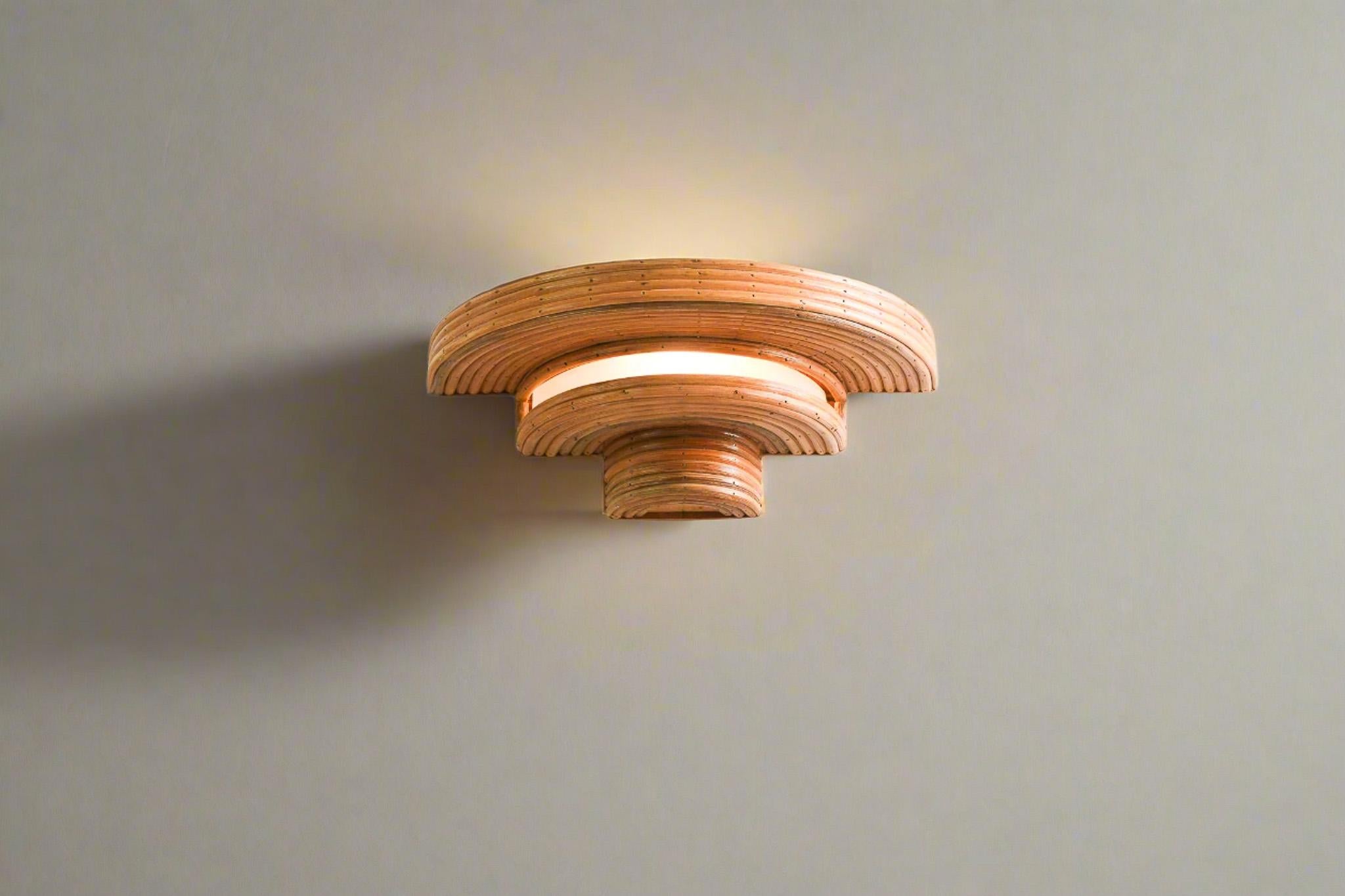 Combinando elementos del pasado y del futuro, esta lámpara de pared muestra una fusión única de estilos de la Era Espacial y el Art Decó. Fabricada con ratán orgánico, sus capas curvadas crean una atmósfera suave y radiante que resalta su calidez