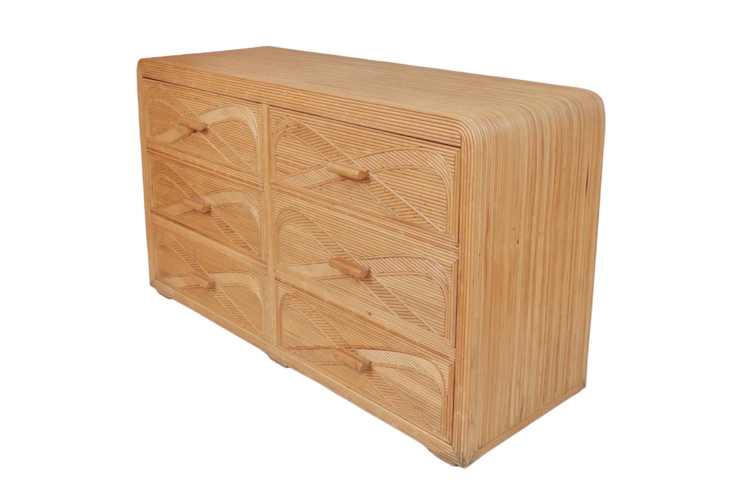 A pencil reed waterfall dresser. Waterfall edges at each side give the piece gentle curves around six drawers that open with wooden bar handles. Drawer fronts are decorated with inlaid pencil wood bows that cross at the handles. Raised on wooden