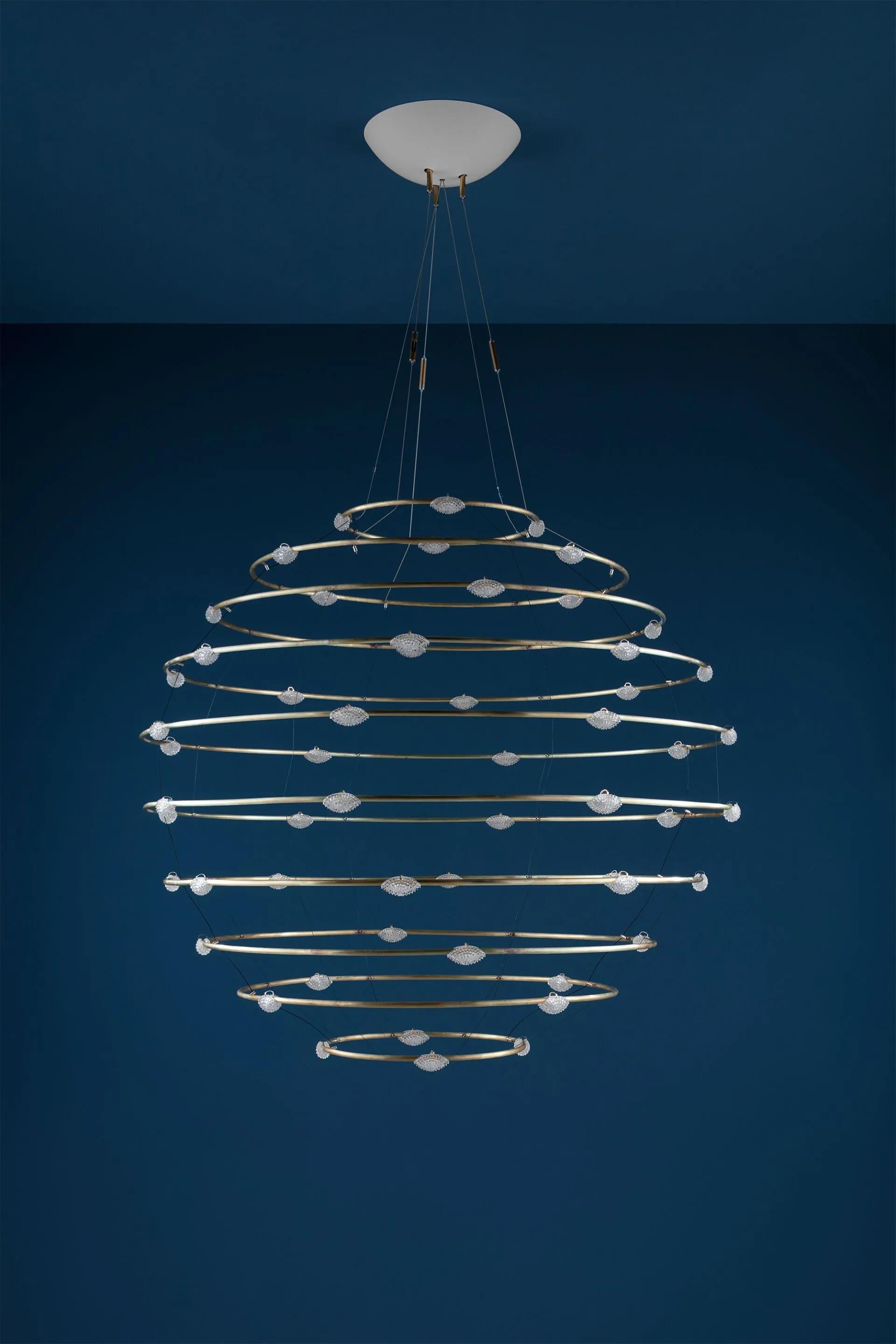 El Petits Bijoux PB56, diseñado por Enzo Catellani, representa un movimiento estático: la ondulación congelada de una piedra que se deja caer en el agua, formando ondas concéntricas que irradian a través del espacio. Esta lámpara colgante continúa