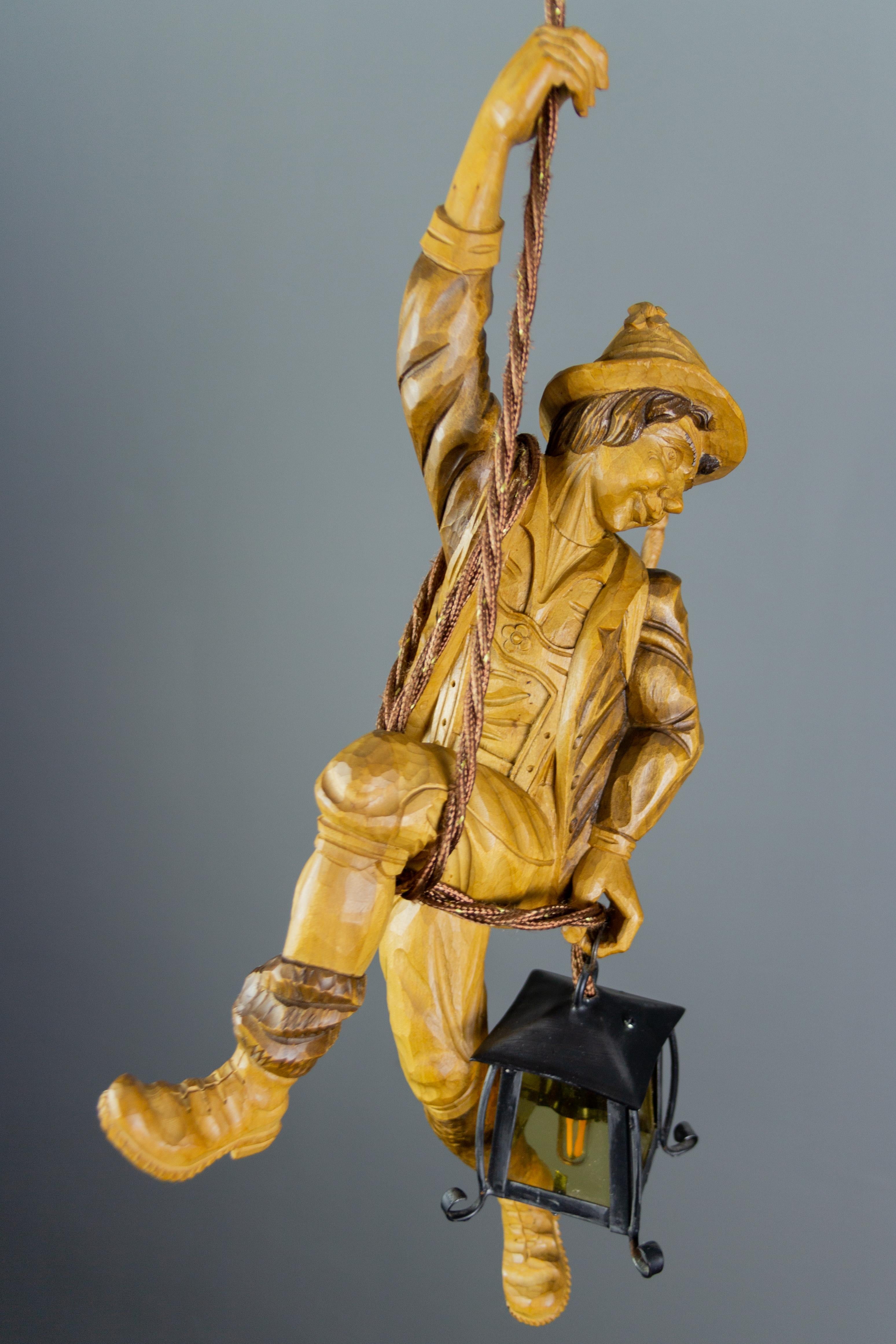 Wonderful German figural pendant lamp features a hand carved figure of a mountain climber in natural brown wood tones. The detailed carved wooden mountaineer with a backpack is holding onto a rope and holding a metal lantern with yellow glass in one