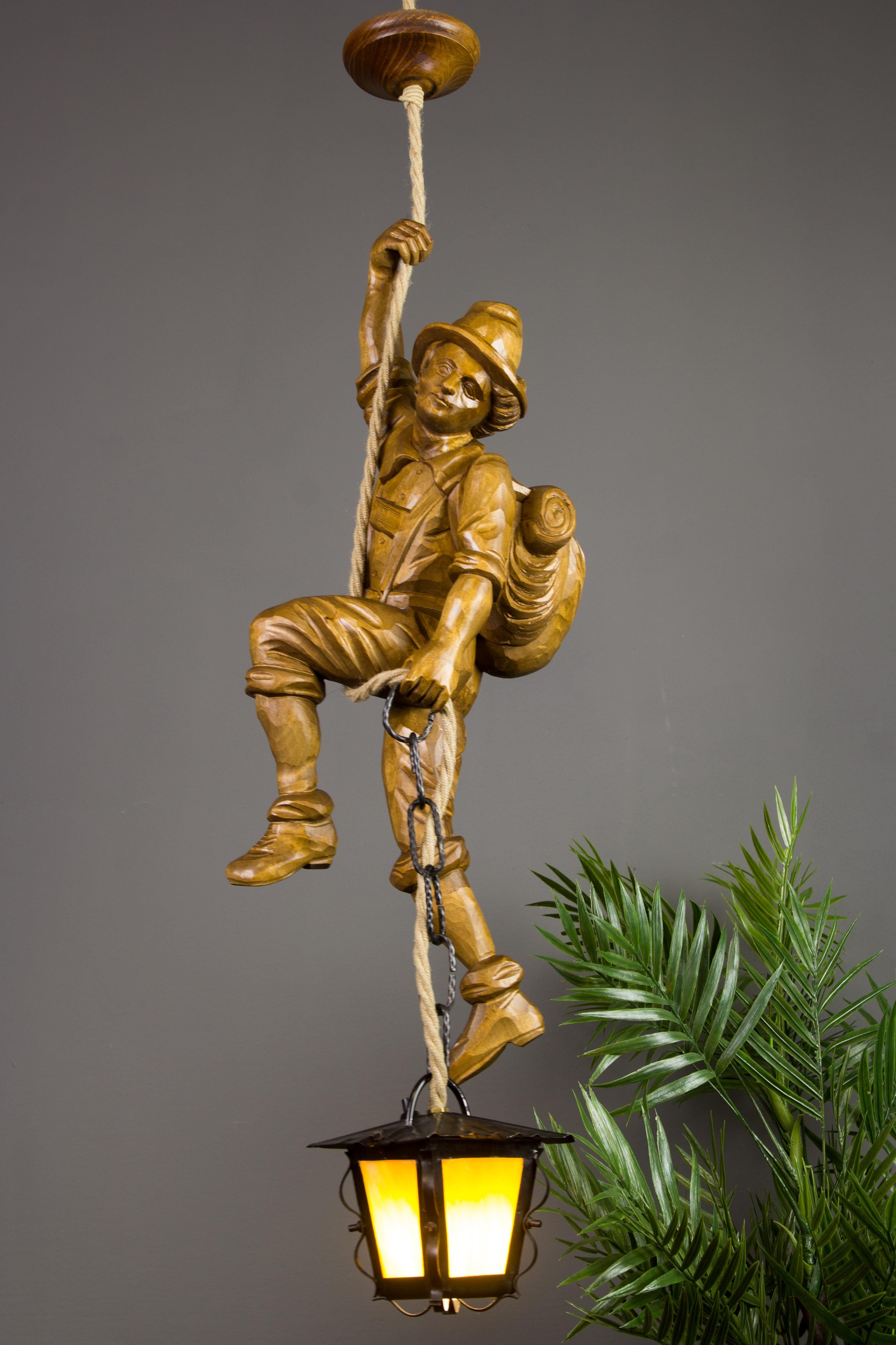 Cette magnifique lampe suspendue figurative de la Forêt Noire allemande présente une figure d'alpiniste sculptée à la main. L'alpiniste en bois sculpté avec un sac à dos s'accroche à une corde et tient dans une main une lanterne en métal avec un