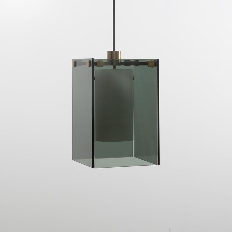 Hanging light made of four dark grey glass sheets held in place by an upper structure in the shape of a cross made of patinated brass. 
A white bulb cover hangs in the centre of the lamp to enable the light to be released in a subtle way. 
 
The