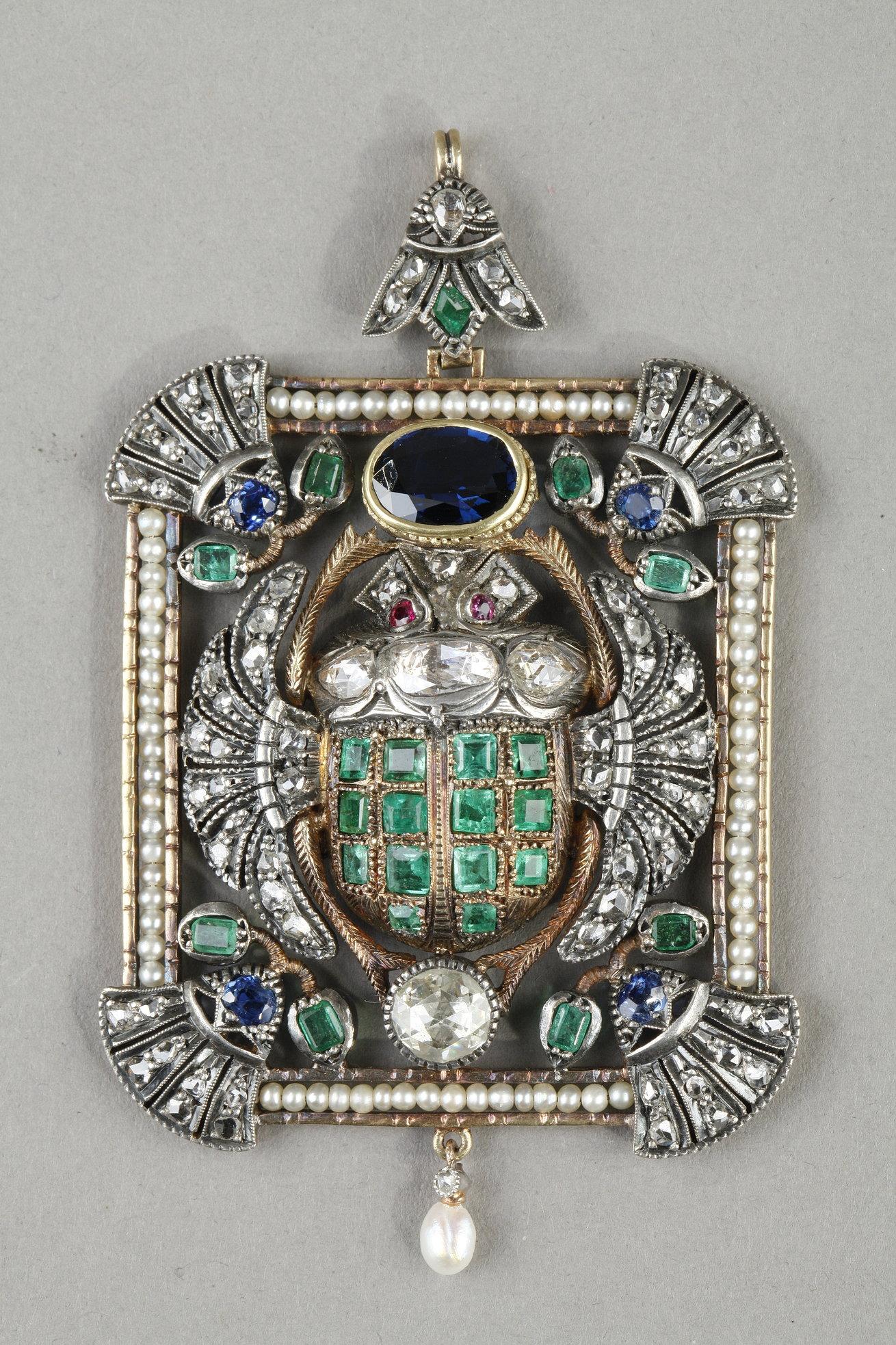 Rectangular gold and silver pendant depicting a winged beetle in a fine pearl frame, the corners of which are decorated with lotus leaf motifs. The beetle is entirely set with sapphire, emerald and diamond stones. It is topped by a cabochon-mounted