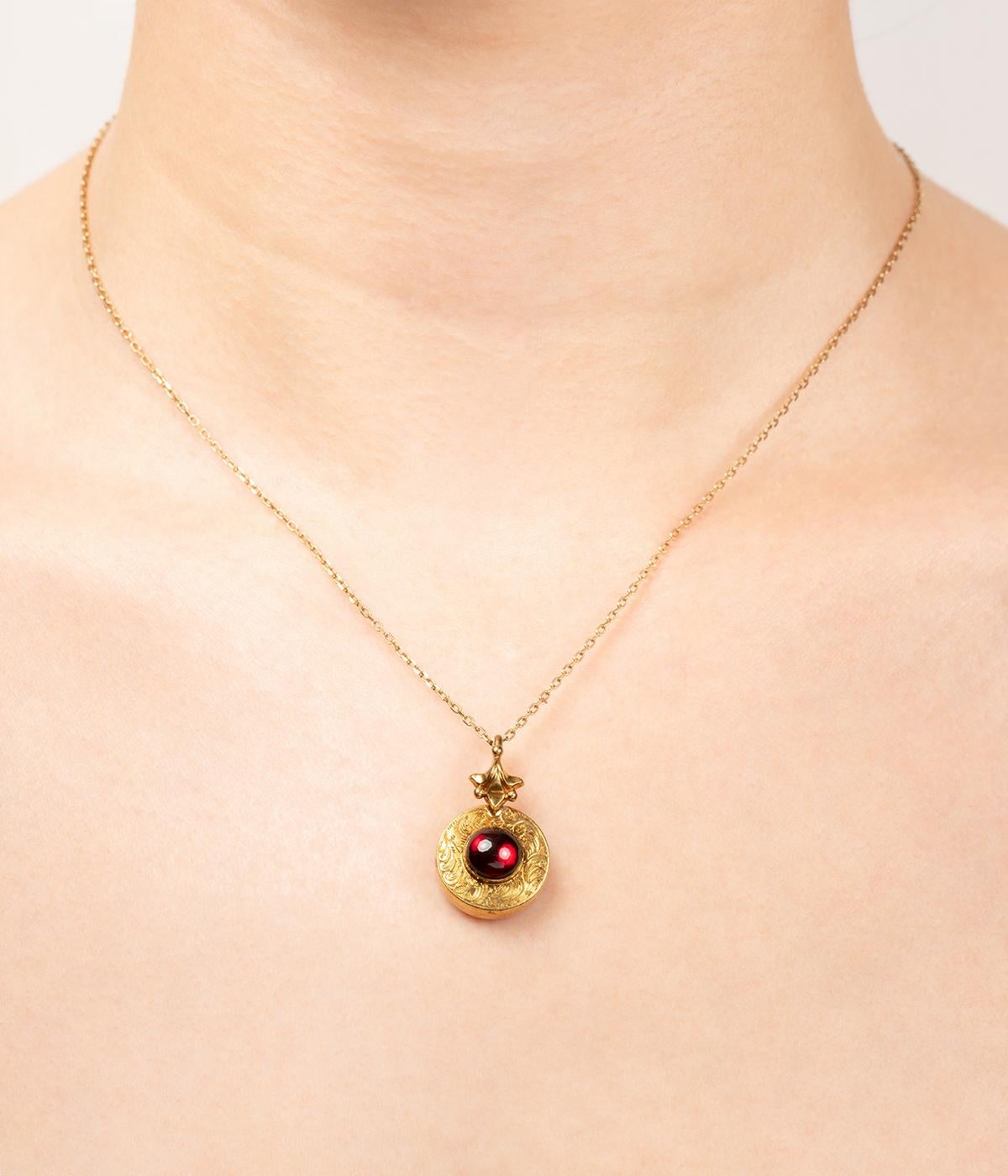 Pendentif porte-souvenir de la fin du 19ème siècle. De forme ronde, il est en métal plaqué or et orné d'un cabochon de grenat en serti clos au centre d'un décor finement ciselé de rinceaux feuillagés. La bélière en or 14 carats (585 millièmes)