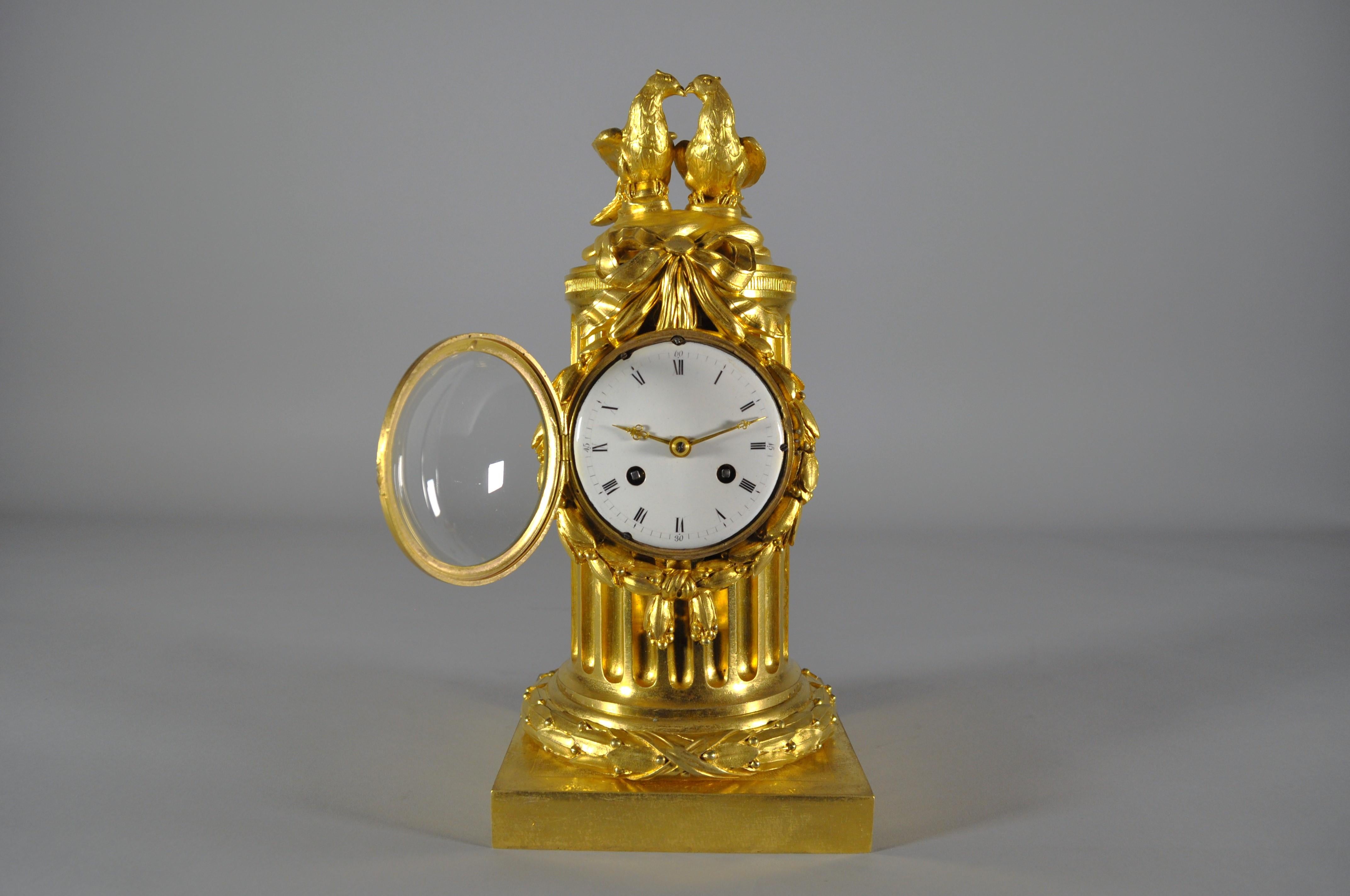 Antique French gilt bronze clock depicting the trunk of a fluted column with a pair of doves resting on top and decorated with a bow and festoon framing the dial. 
White enamel dial with black enamel Roman numerals. Golden hands. 
Gilt brass
