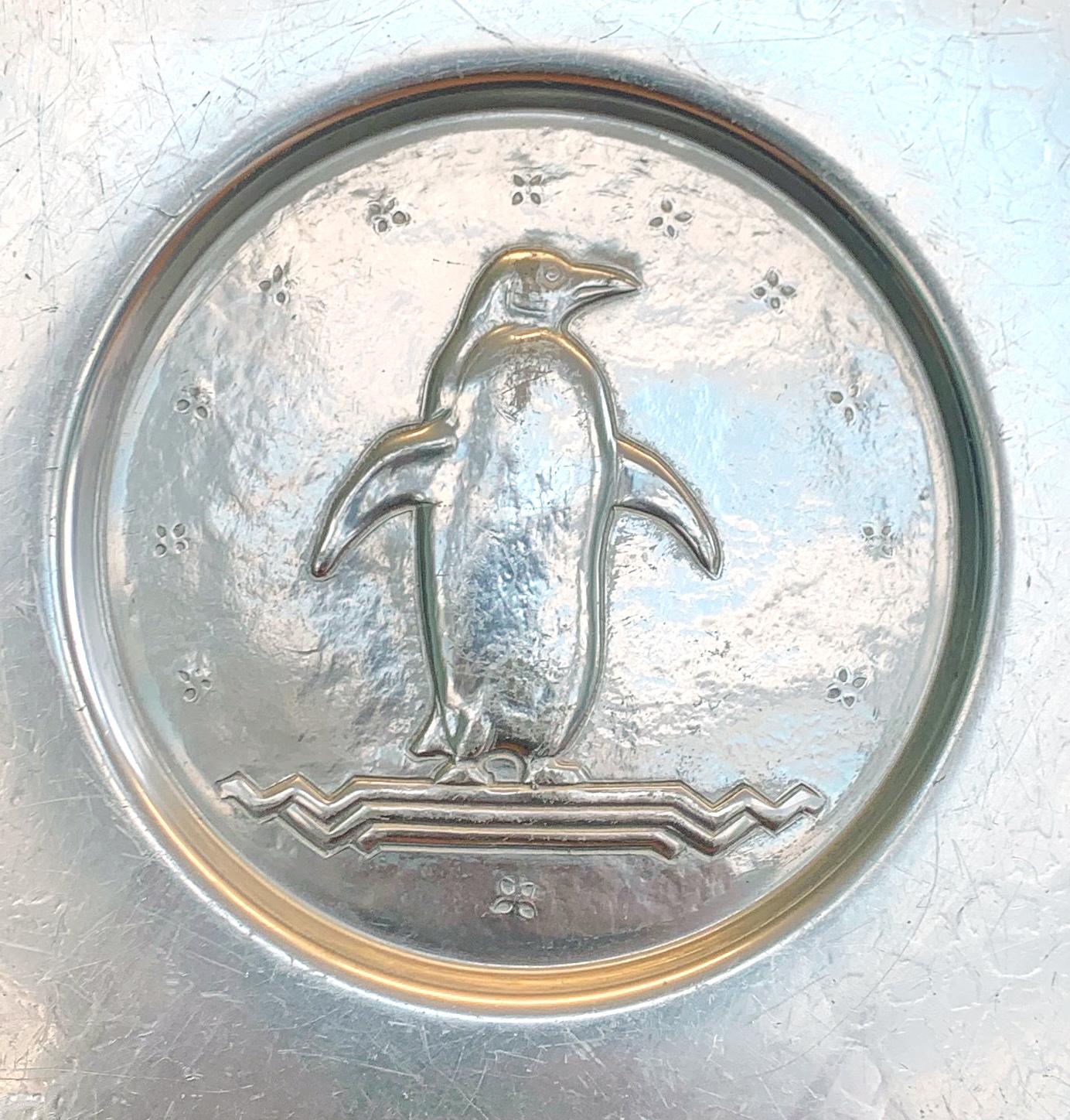 Striking and rare, this glinting charger featuring a penguin standing atop an Art Deco ledge is composed of a silvery aluminum finished in a subtly iridescent glaze. This piece, along with an array of chargers and bowls in the same series, was