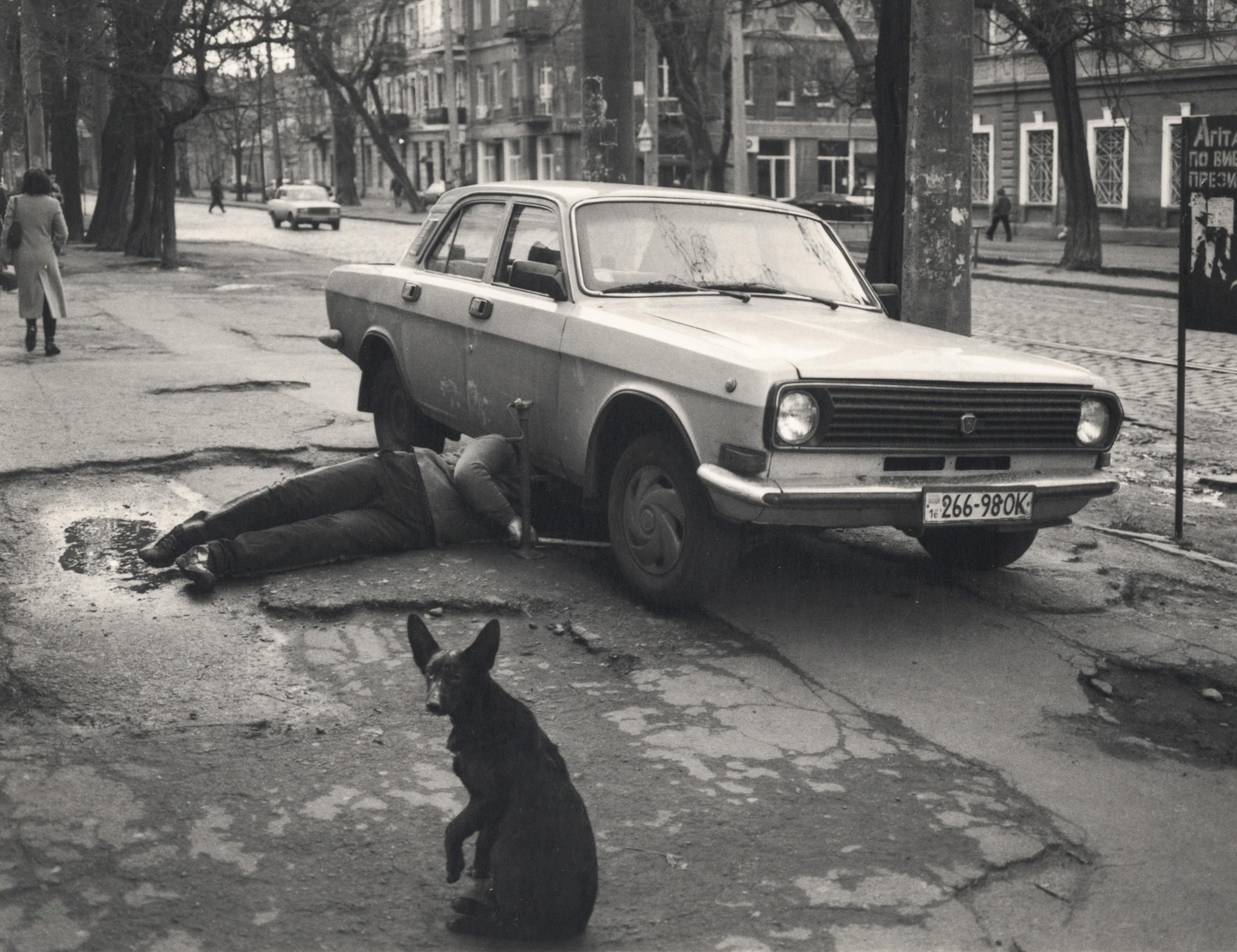 Pentti Sammallahti Black and White Photograph - Odessa, Ukraine (Man repairing vehicle on sidewalk & Black dog)