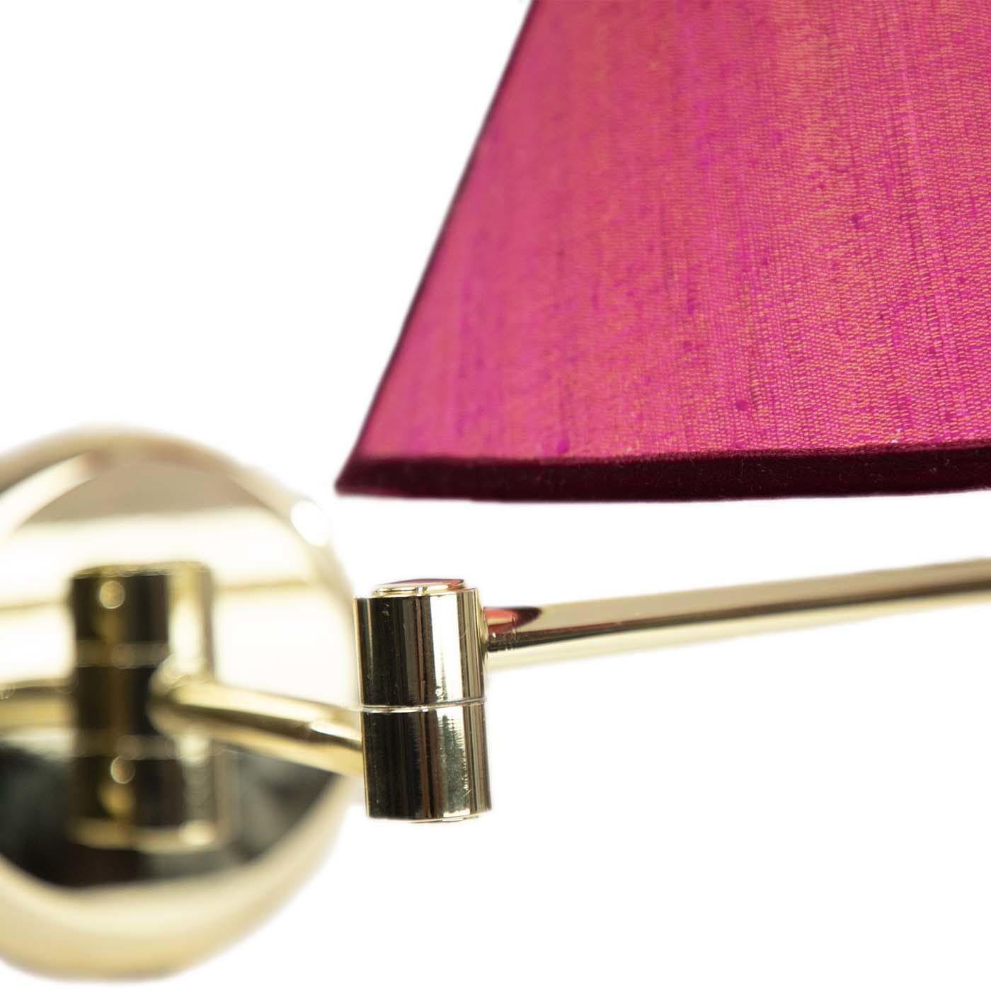 The Pepita wall light boasts a gold metal base, equipped with a practical swivel arm and chain switch mechanism. Its timeless conical shade is upholstered in vibrant pink raw silk, featuring burgundy velvet trims for a refined finish. A seamless