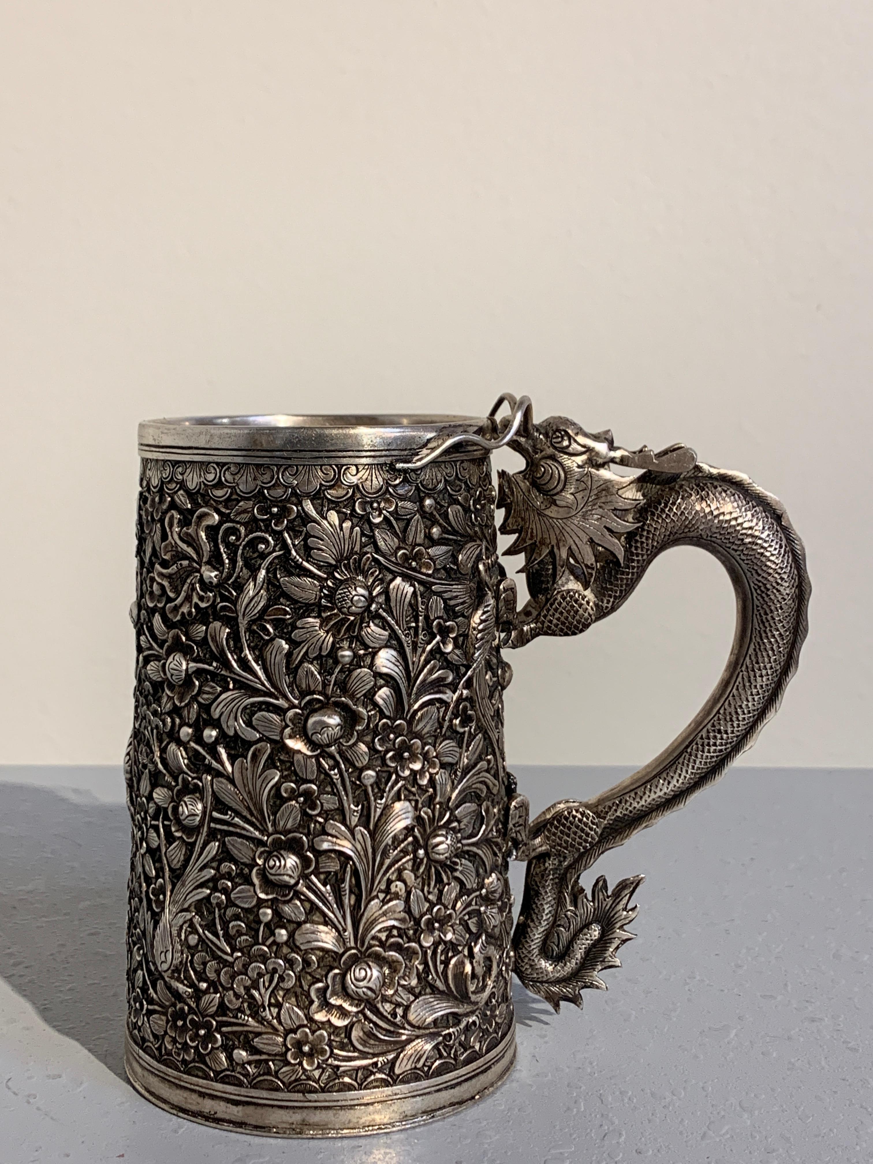 A heavy and well cast Chinese export silver tankard with a dragon handle, made of the Southeast Asian market, mid-late 19th century, China.

The double walled mug or tankard featuring a dense and exuberant scene of long tailed birds amongst a lush