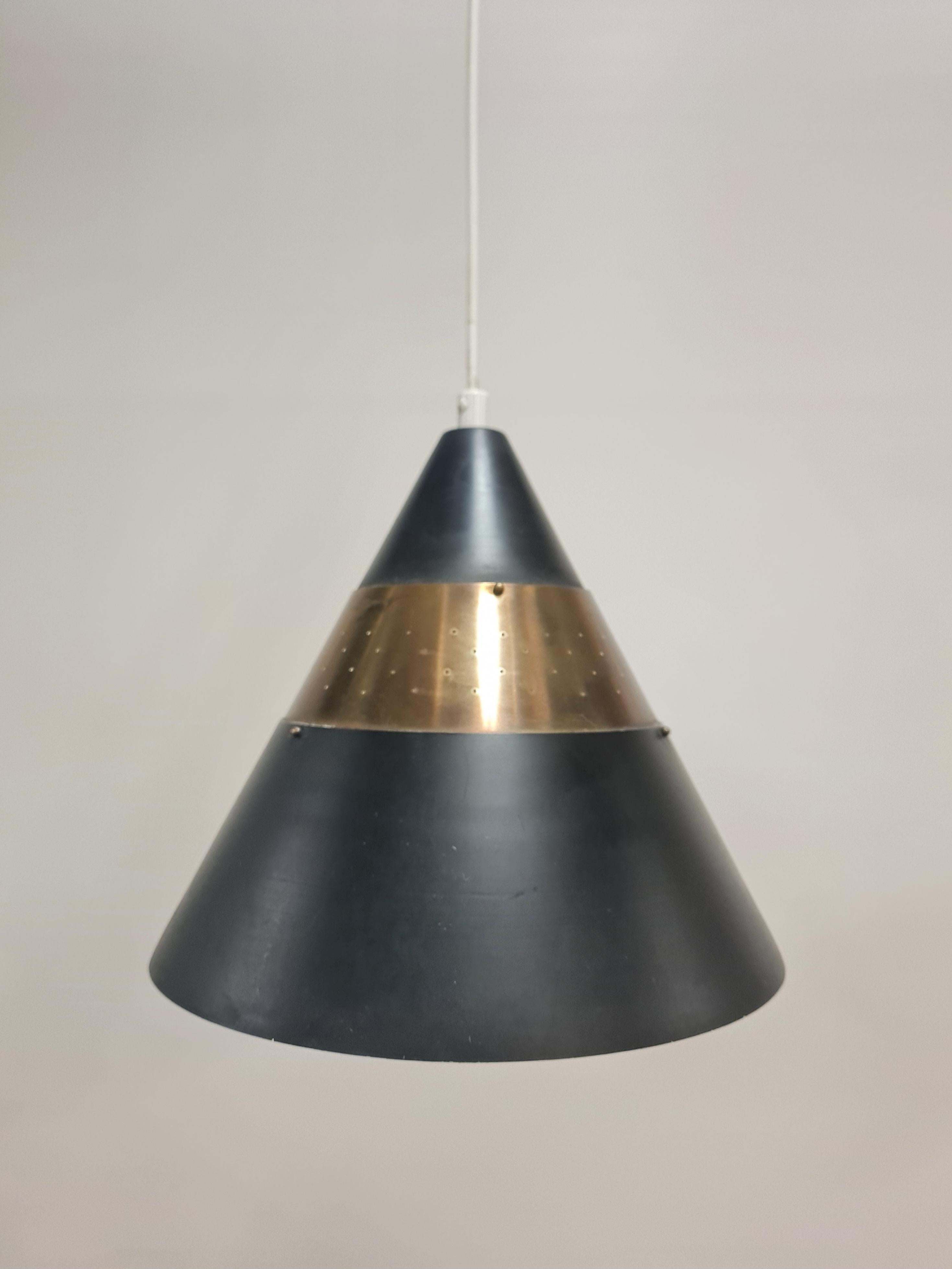 Un pendente da soffitto a forma di cono, perforato e stratificato, realizzato da Maria Lindeman per Idman. La lampada è composta da tre strati di metallo, due in grigio scuro e uno in ottone perforato. La lampada è nelle sue condizioni originali e
