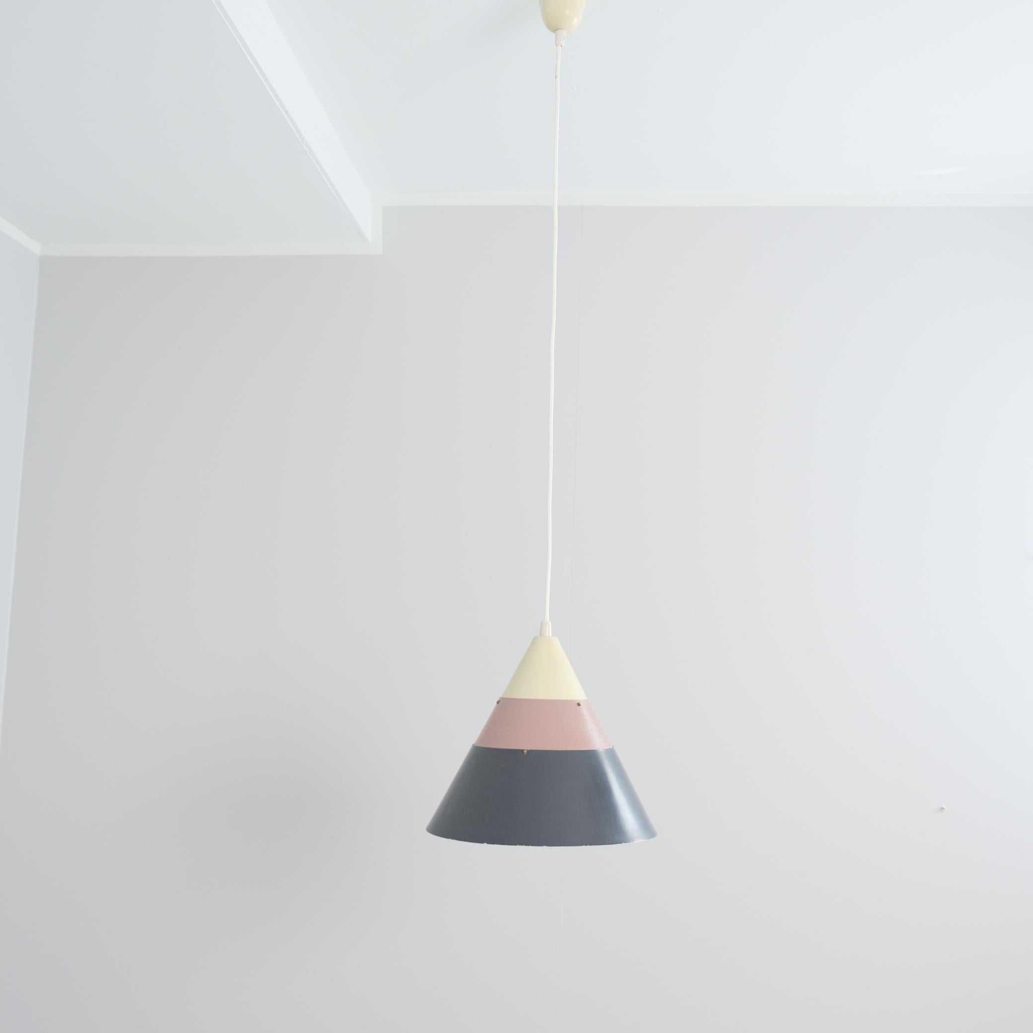 Un pendente da soffitto a forma di cono, perforato e stratificato, realizzato da Maria Lindeman per Idman. La lampada è composta da tre strati di metallo di colori diversi che vanno dal grigio scuro al beige brik e al beige chiaro. La lampada è