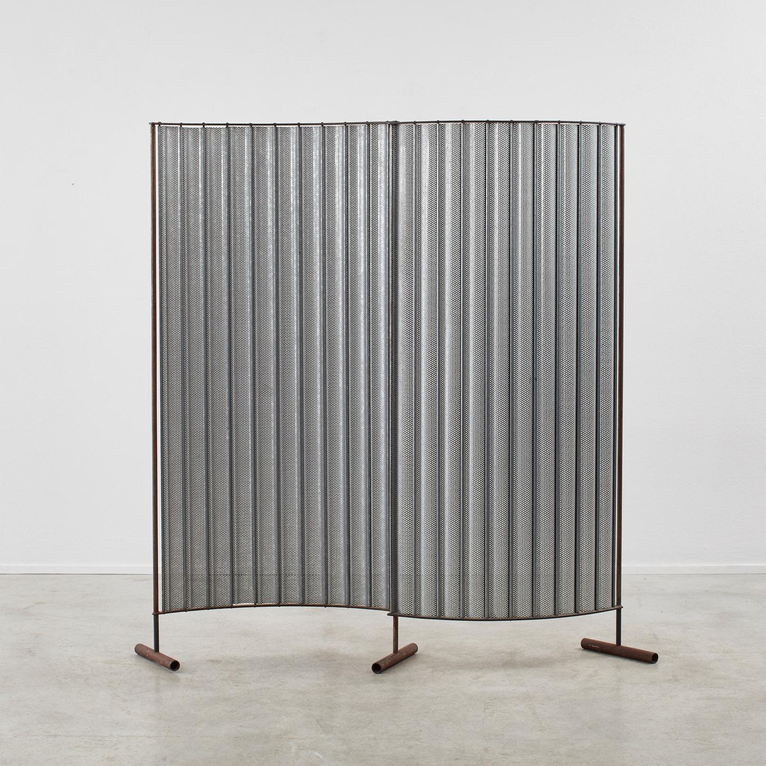 A corrugated steel screen stands atop three wooden legs. Its sinuous arc and hundreds of delicate perforations strike a pleasing balance to its industrial choice of metal. 

This screen carries all the stylistic hallmarks of a modernist design, and