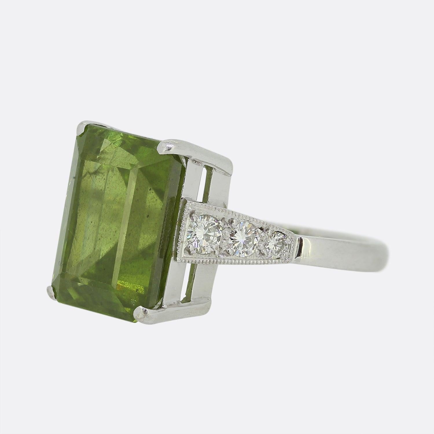 Here we have an 18ct white gold peridot and diamond cocktail ring. The centre stone is a large rectangular shaped peridot possessing an intense dark green hue. This focal gemstone is flanked on either shoulder by a trio of milgrain set round