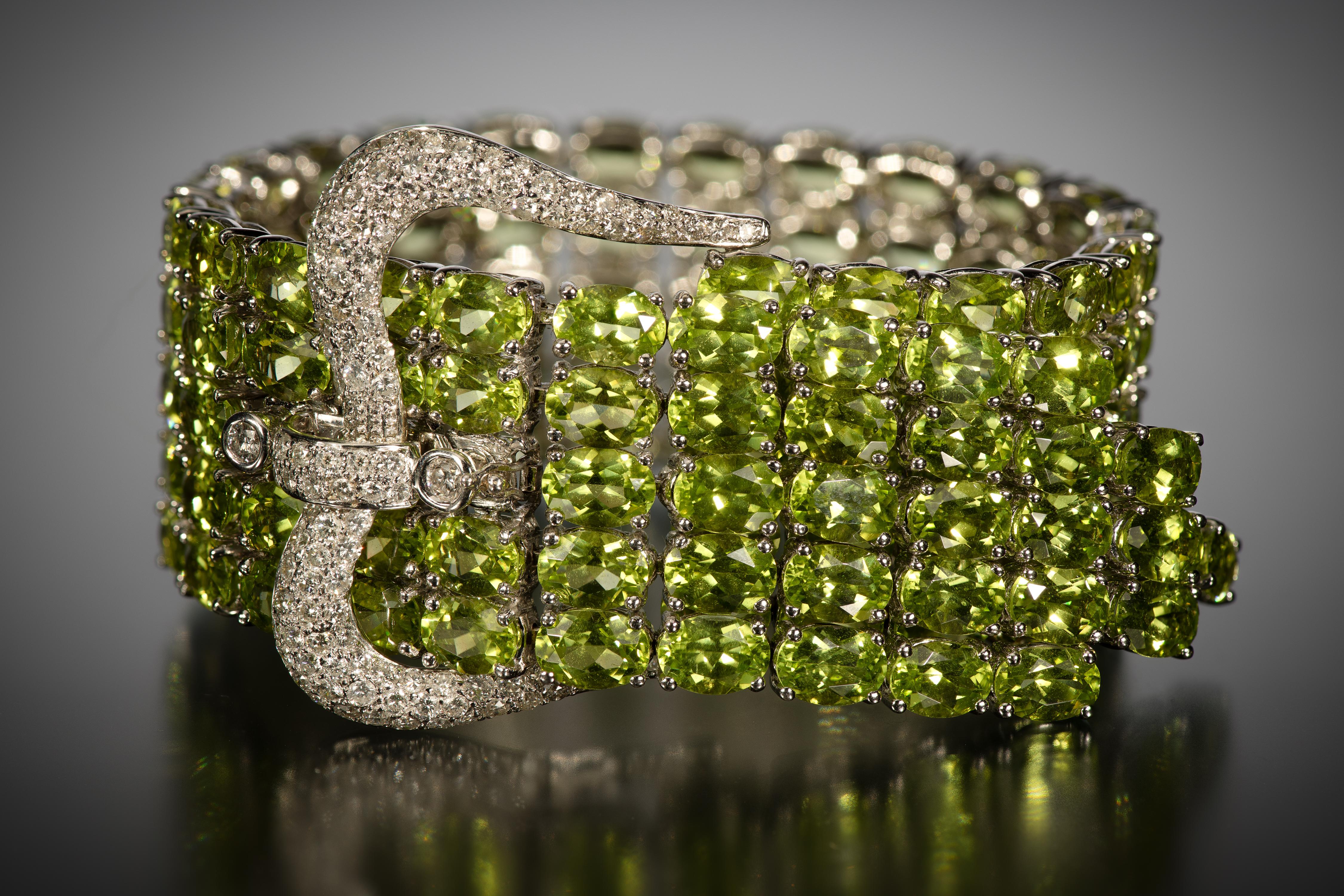 A wide strap of bright green peridots wraps around the wrist, ending in a flourish of peridot fringe. The bracelet is secured by a crown-shaped buckle set with over 2 carats of bright white diamonds. The peridots are the most desirable shade of