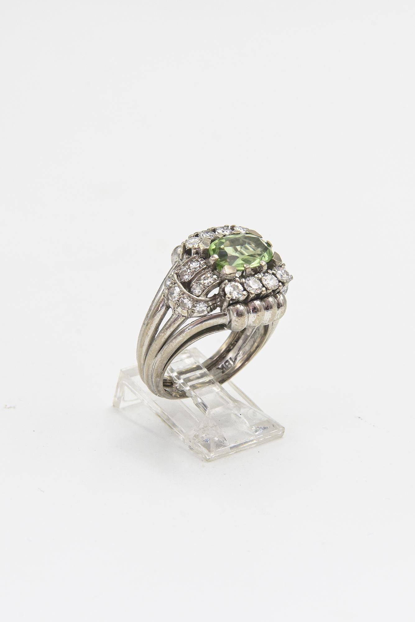 Highly stylized mid 20th century 18k white gold cocktail ring featuring an oval peridot center stone.  On the top and bottom of the peridot are 4 prong set diamonds above which is a 4 section gold roll. On the shoulders there is a diamond garland