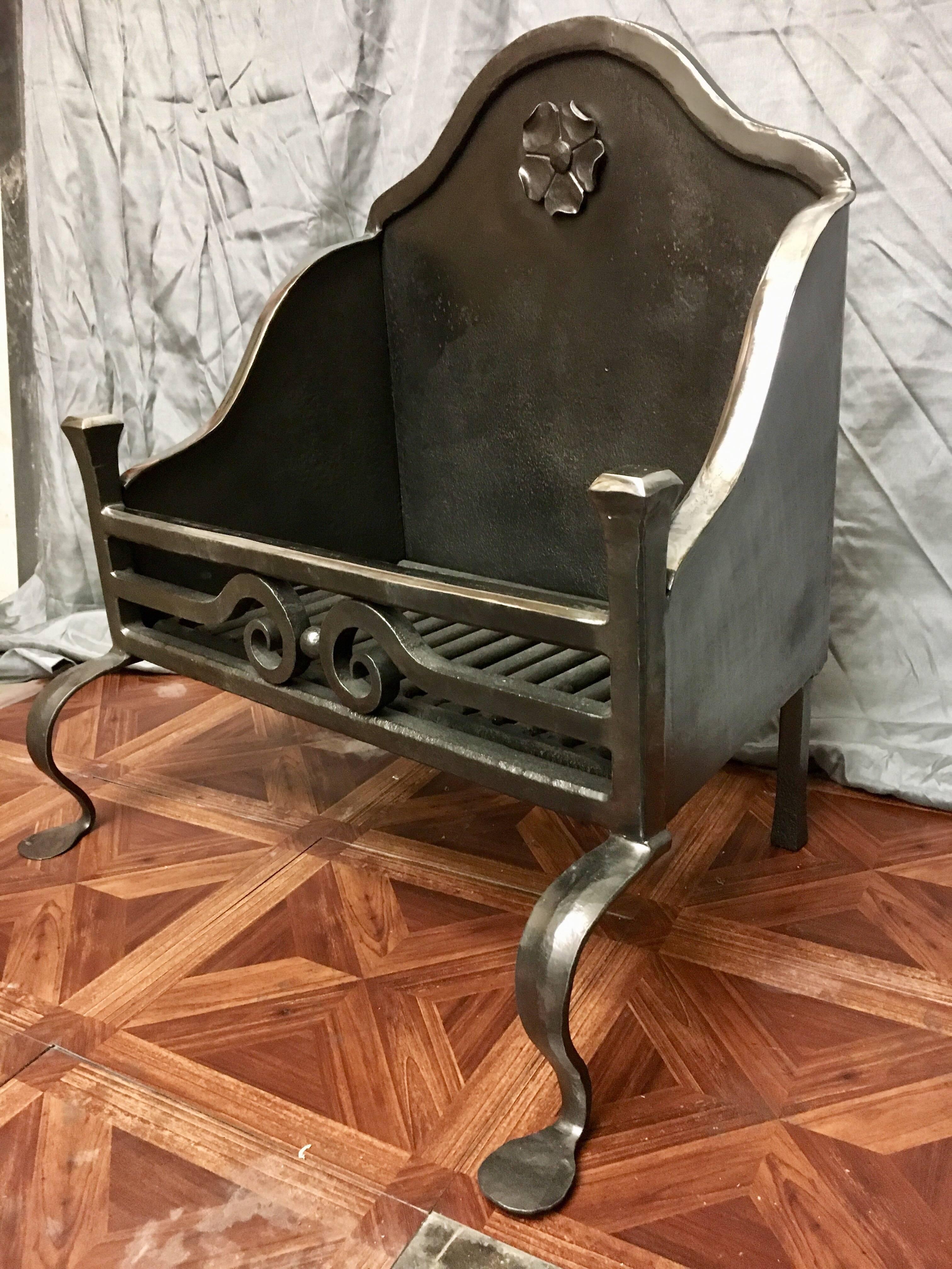 A simple, early 20th century wrought Iron fire basket with a serpentine shaped fire back with hammered lip and a central stylized flower embellishment. Tapered lipped side panels connect to a three bar fire front with central handed pigs tail