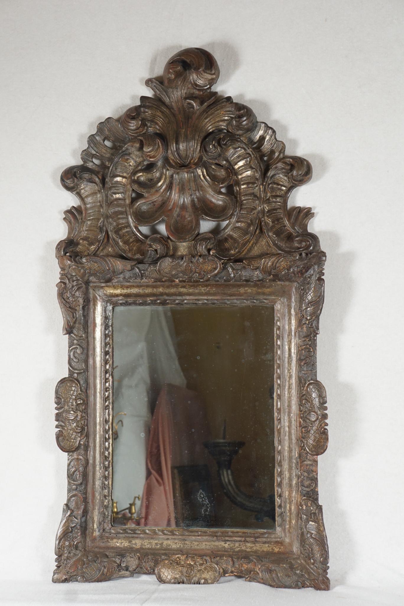 This fine old mirror from circa 1740 is created in carved wood, gessoed and silver gilded. Its bold and heavy frame is centered at the top by a dramatic openwork crest consisting of large carved s and c scroll elements with a dramatic stylized shell