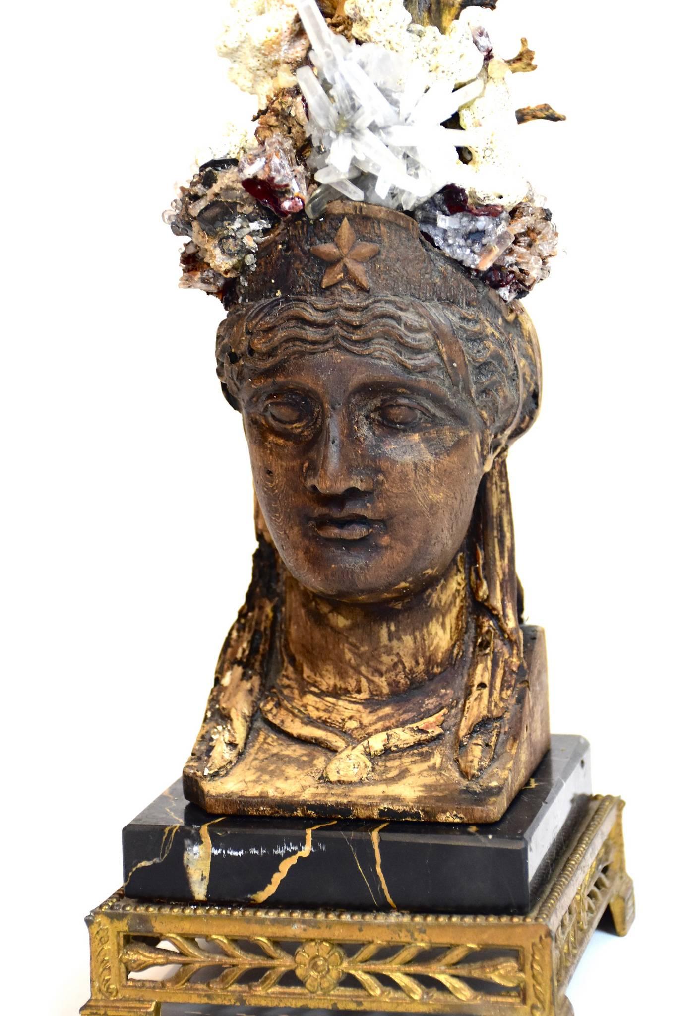 A 200 year old Empire carved bust fragment is adorned with an organic blooming crown to include a giant black coral sea fan, corals, andradite garnets, hemimorphite and quartz specimens. The base is a 19th century Michelangelo marble slab sitting on