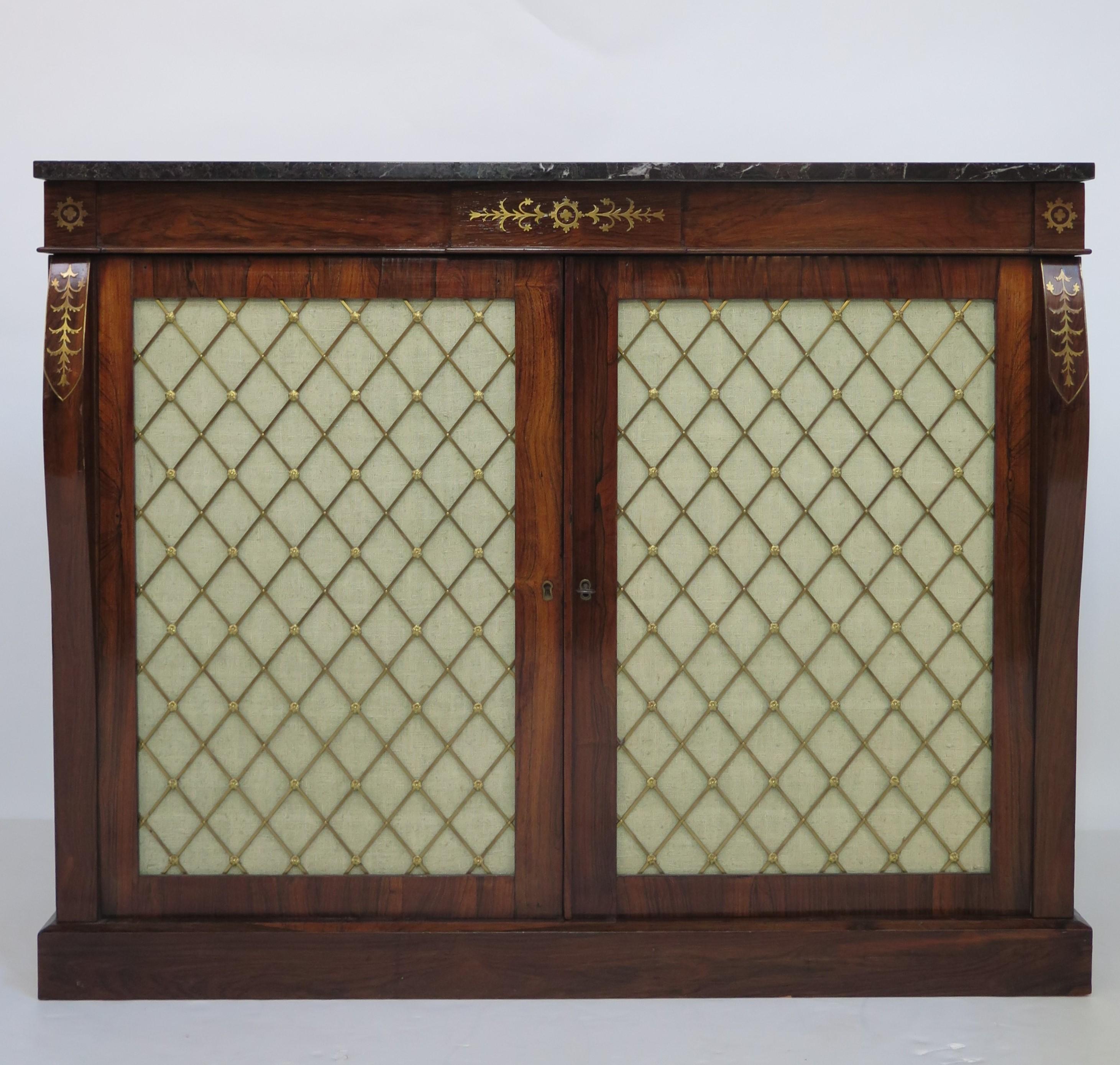 An elegant period English Regency cabinet chiffonier of rosewood. Curving pilasters with floral brass inlay at sides of two brass grillwork doors with fabric liner that open to reveal a cupboard with a central shelf. Black marble top with white