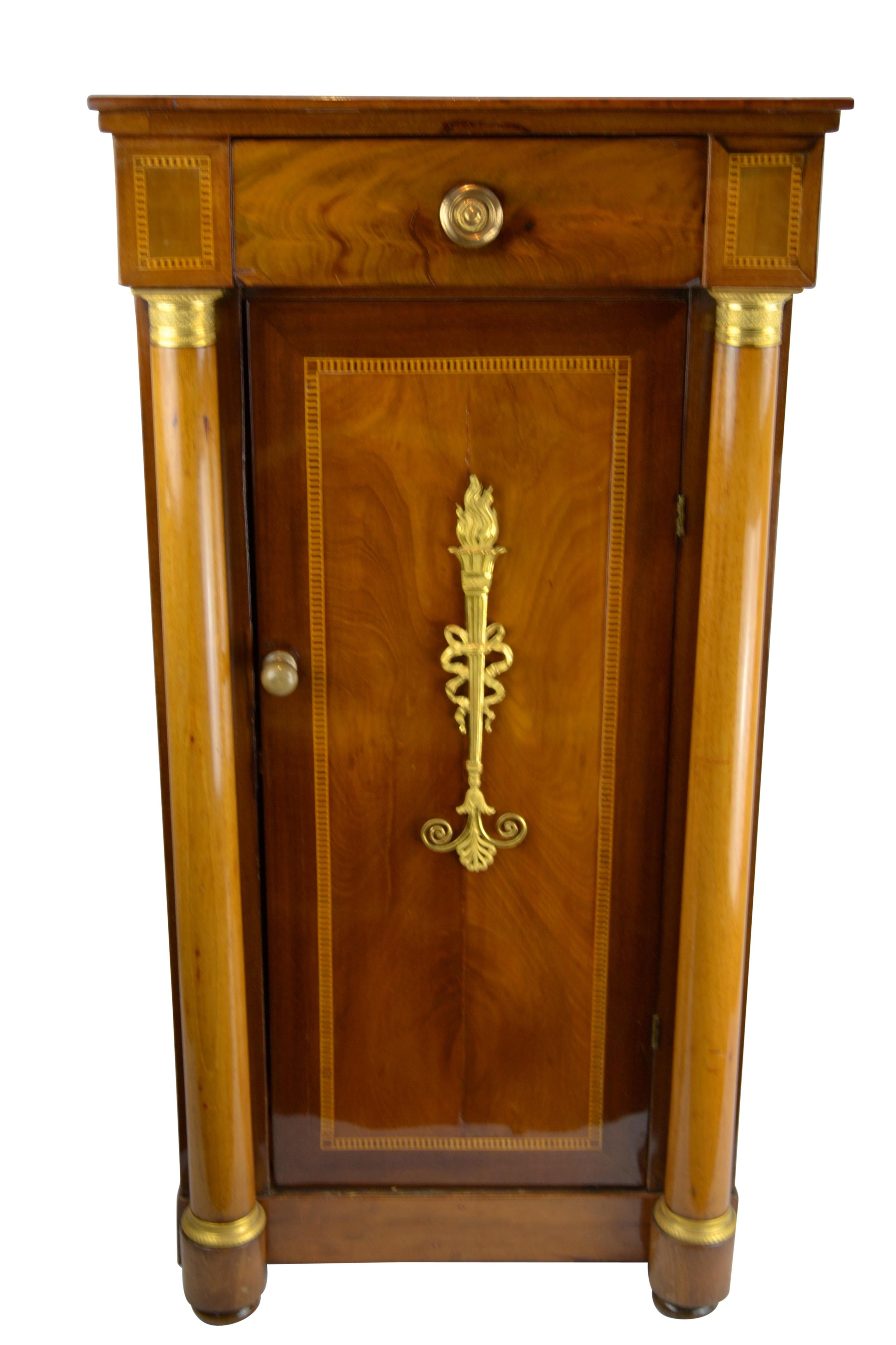 An Empire bedside table in beautifully faded mahogany and inlaid with various other exotic woods to the front and sides. The front door is framed in circular columns with gilt moldings to the top and bottom. The front door is decorated with a fine