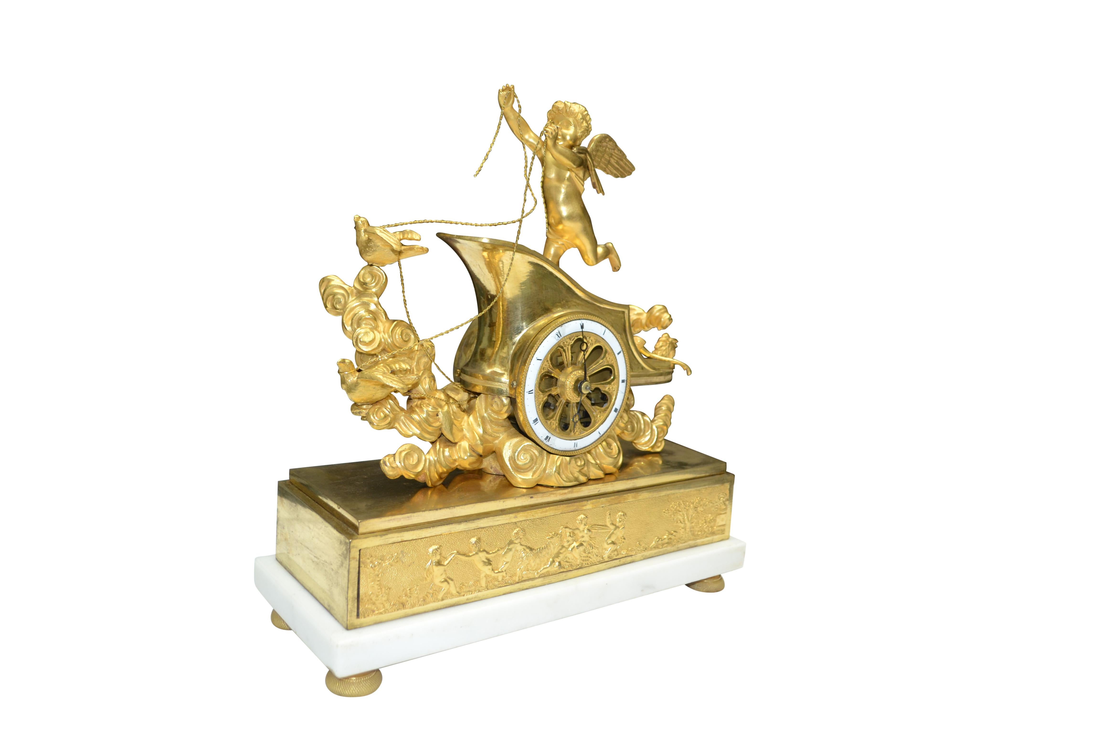 The clock features a winged cupid standing on his chariot drawn by two doves and supported on clouds; cupid holds the reins to two doves which appear to be guiding the chariot. The open 'wheel' of the chariot contains the outer enamel clock dial