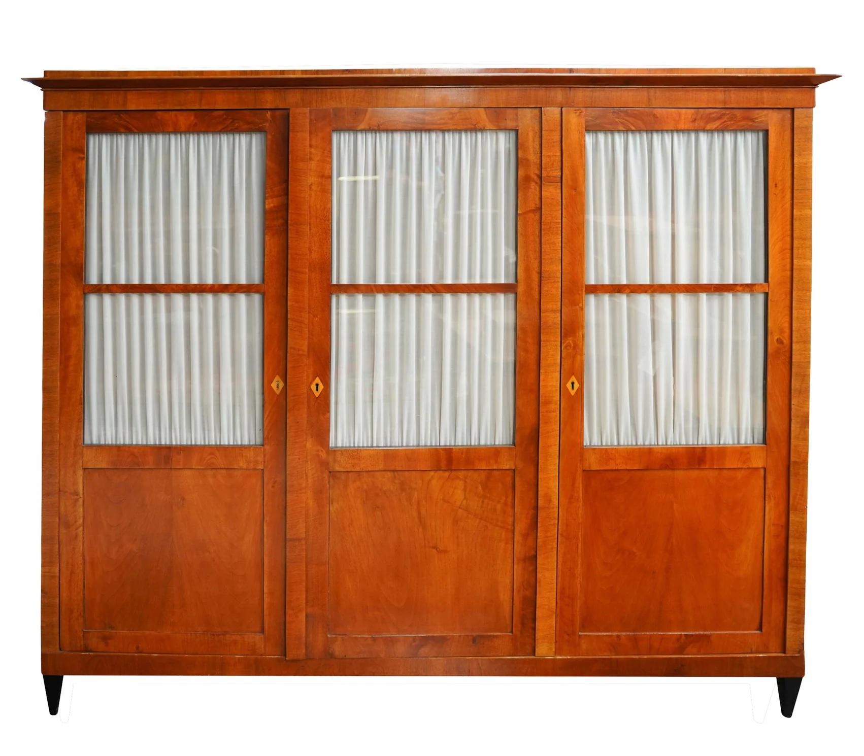 Period Antique Early 19th Century (Circa 1820) Fruitwood German Biedermeier Three Door Glazed Cabinet Rising on Four Ebonized Tapered Legs. The three divided lite glazed doors with privacy curtains open to 3 vertical compartments with removable,