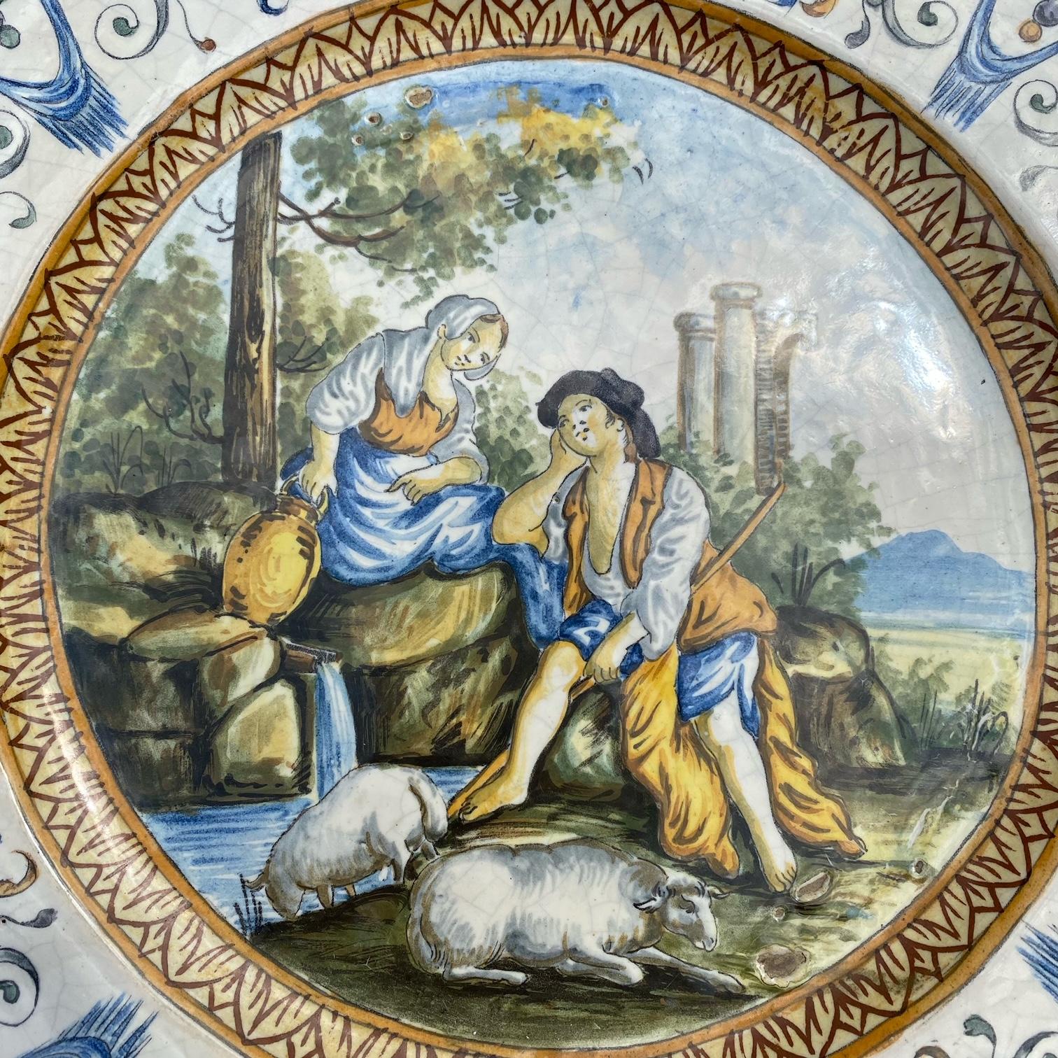 This beautifully hand painted Italian Majolica plate is made of tin glazed earthenware, from the early 1800s. The mythological scene is one of love, with a shepherd and sheep, and a woman holding an urn. The border of the plate depicts a motif of
