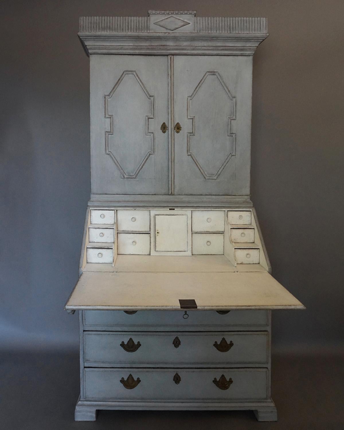 Two part Norwegian secretary, circa 1820, in gray paint with the original locks and brass hardware.

The upper section has a pair of paneled doors opening onto two fixed shelves, all under a reeded cornice which has a central panel with an applied