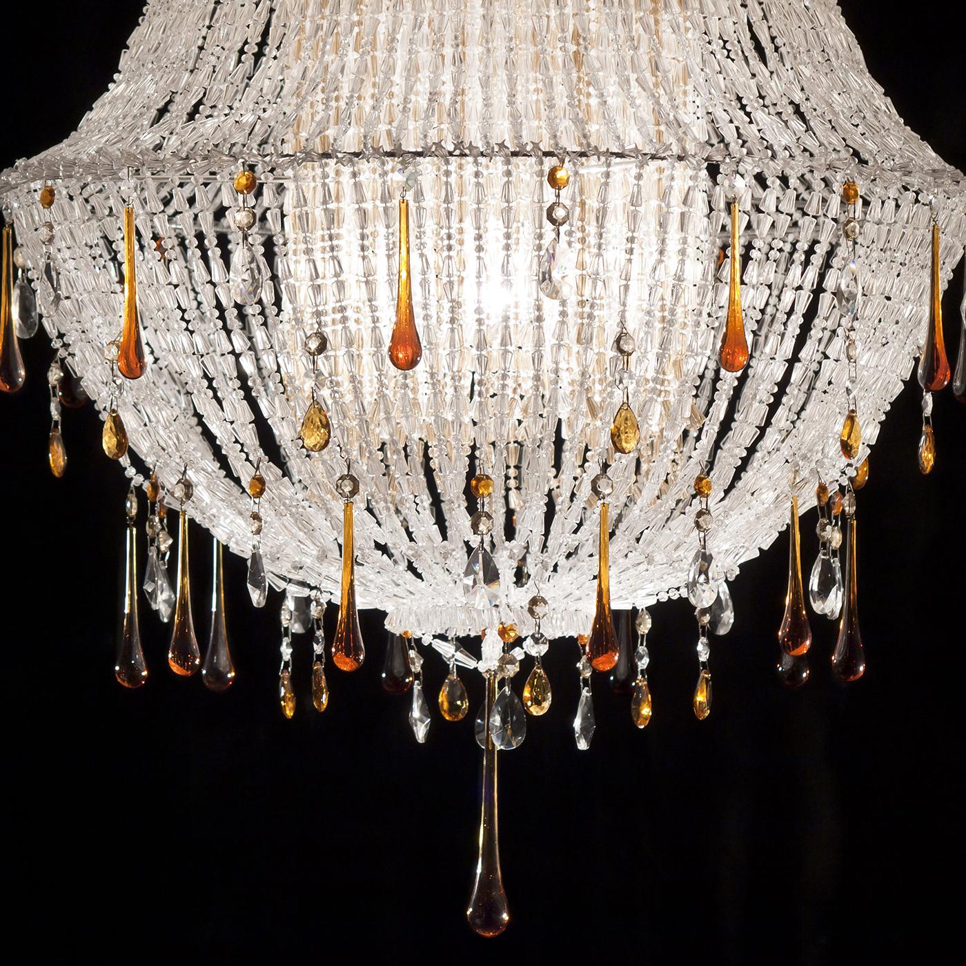 Marked by a flared silhouette, this pendant lamp owes its outstanding charm to the reflecting power of its components, all meticulously hand-tied. Strands made up of transparent acrylic beads mix with amber, teardrop-like crystal, and glass