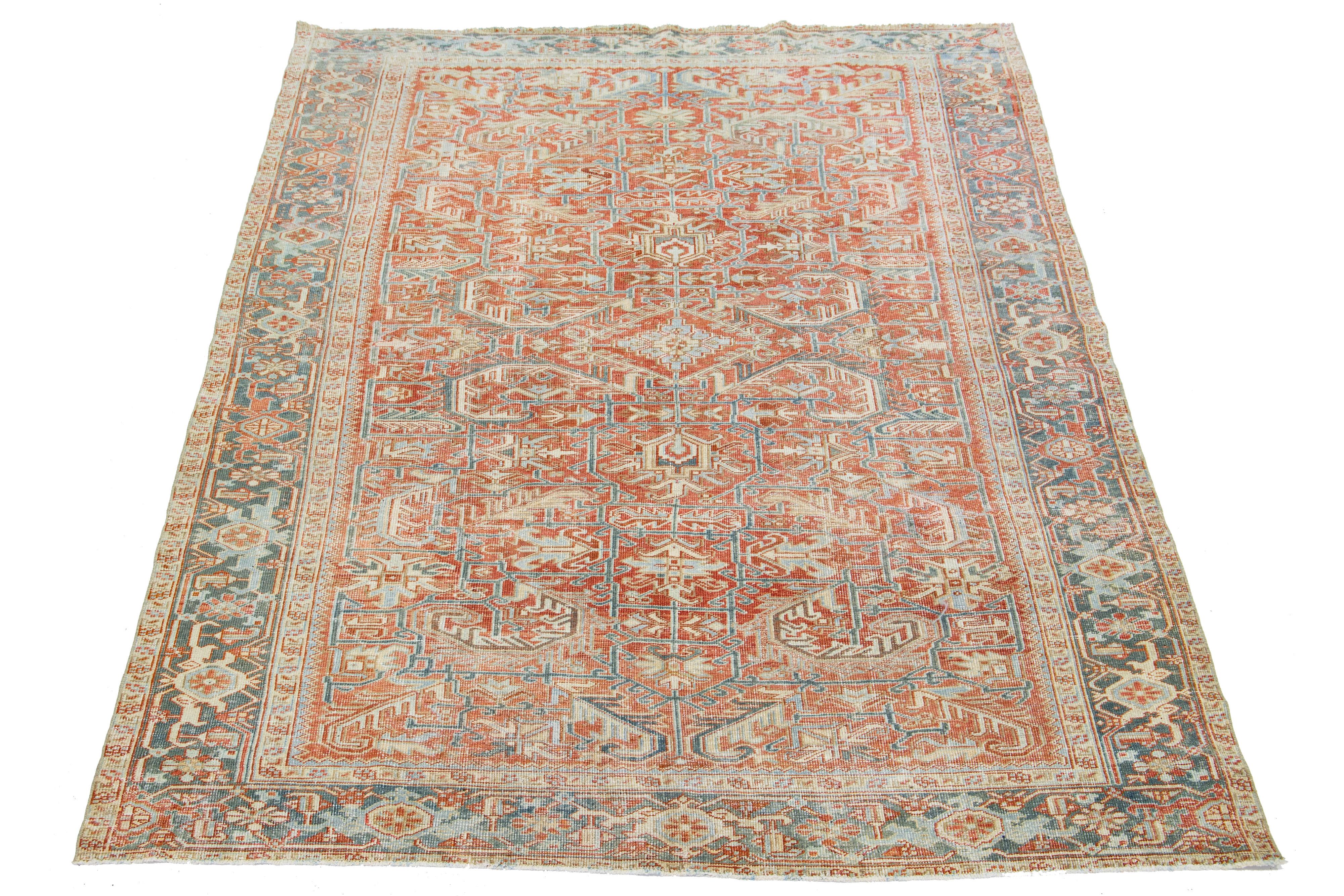This antique Persian Heriz rug showcases an impressive overall design and is crafted with hand-knotted wool. The rust-colored background is a striking backdrop for the captivating geometric floral pattern, embellished with shades of blue, beige, and