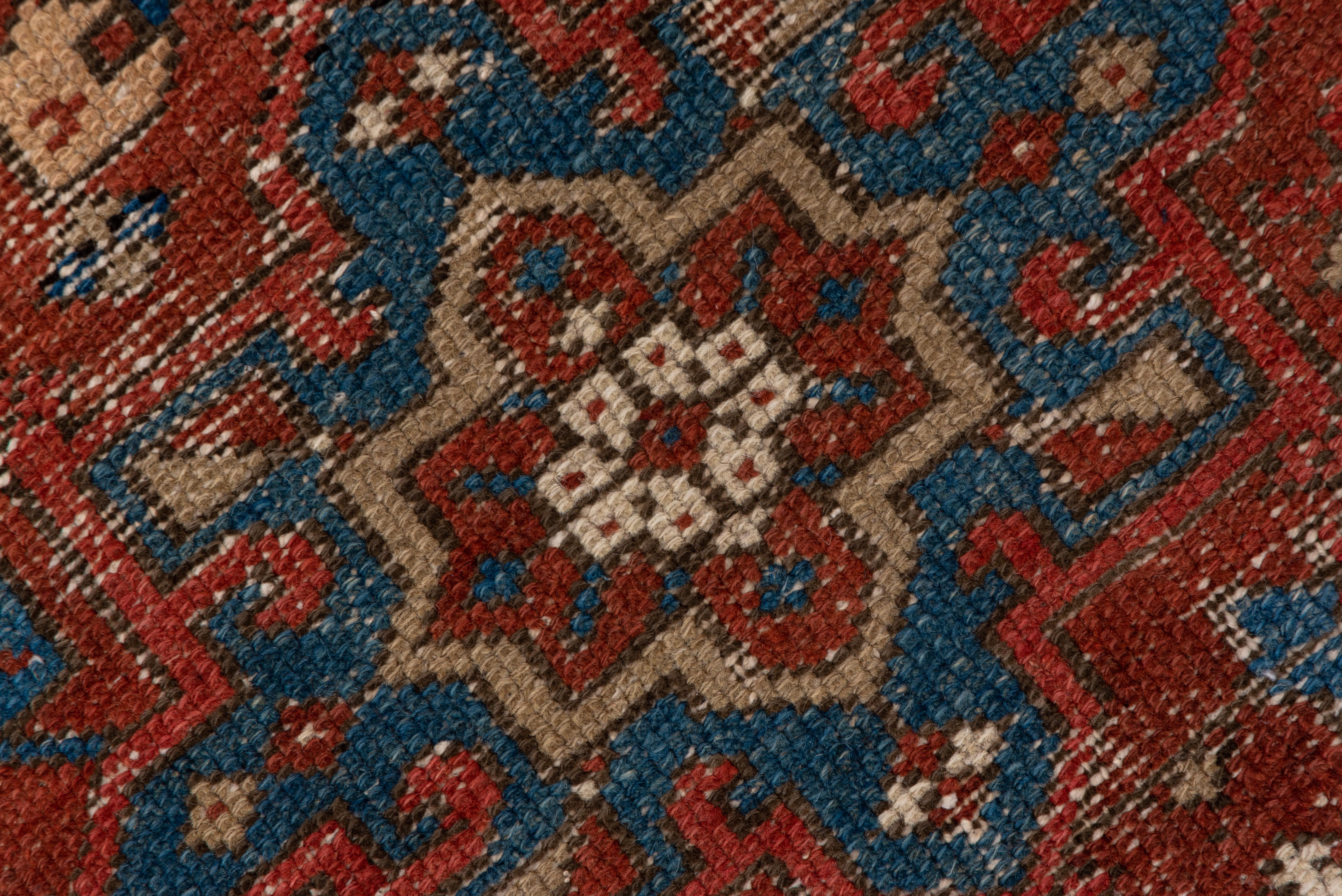 The ketchup red field of this worn, almost distressed NW Persian rustic scatter shows green and buff characteristic octogramme and hooked hexagon medallions. The royal blue border exhibits squared rosettes in ecru, straw, red and green.