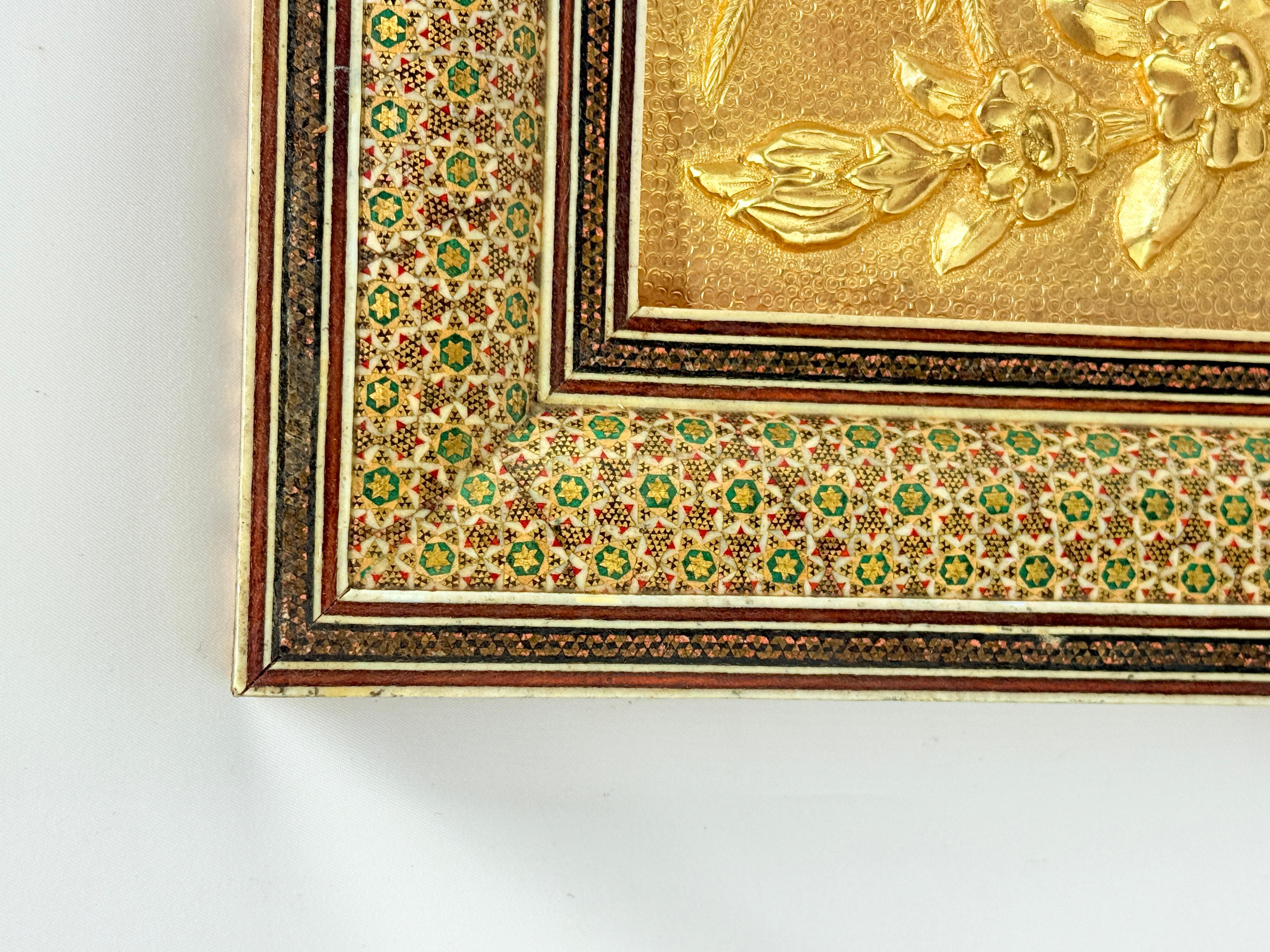 A finely executed Persian decorative panel featuring a raised repoussé and chased scene of birds among blossoming branches, likely crafted from gilt copper or brass and housed in a traditional Khatamkari inlaid wooden frame. The central tableau