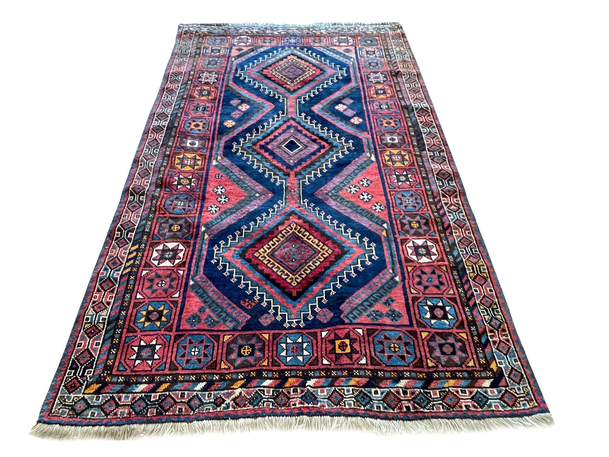 This rug is an authentic rug from Iran, Shiraz. The pile and foundation are wool. The base color is a dark blue with accents of mauve, yellow, blue and brown throughout the rug. The design is geometric with repeated diamond medallion with tribal