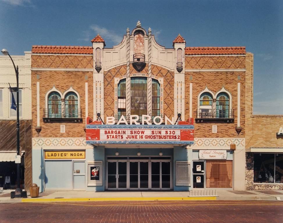 Barron Theater, Pratt, KS von Peter Brown ist ein farbiger Archivpigmentdruck, der die Fassade des Barron Theaters entlang einer Straße zeigt. Das Schild des Kinos wirbt für eine Schnäppchenvorstellung und die Premiere von Ghostbusters 2. Das Kino