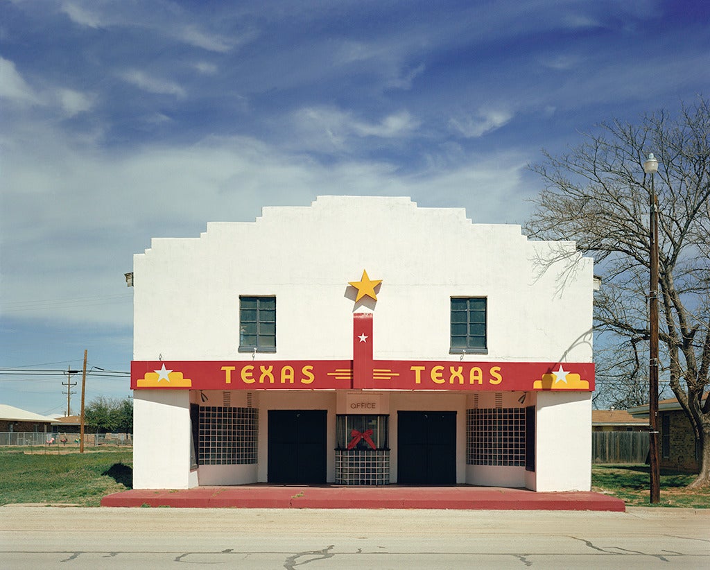 Bronte, Texas von Peter Brown zeigt ein leeres Theater in einer westtexanischen Stadt. Das Theater ist weiß gestrichen, mit Akzenten in leuchtendem Rot und Gelb. Über den Türen des Theaters ist "Texas" gemalt, und zwischen zwei Fenstern prangt ein