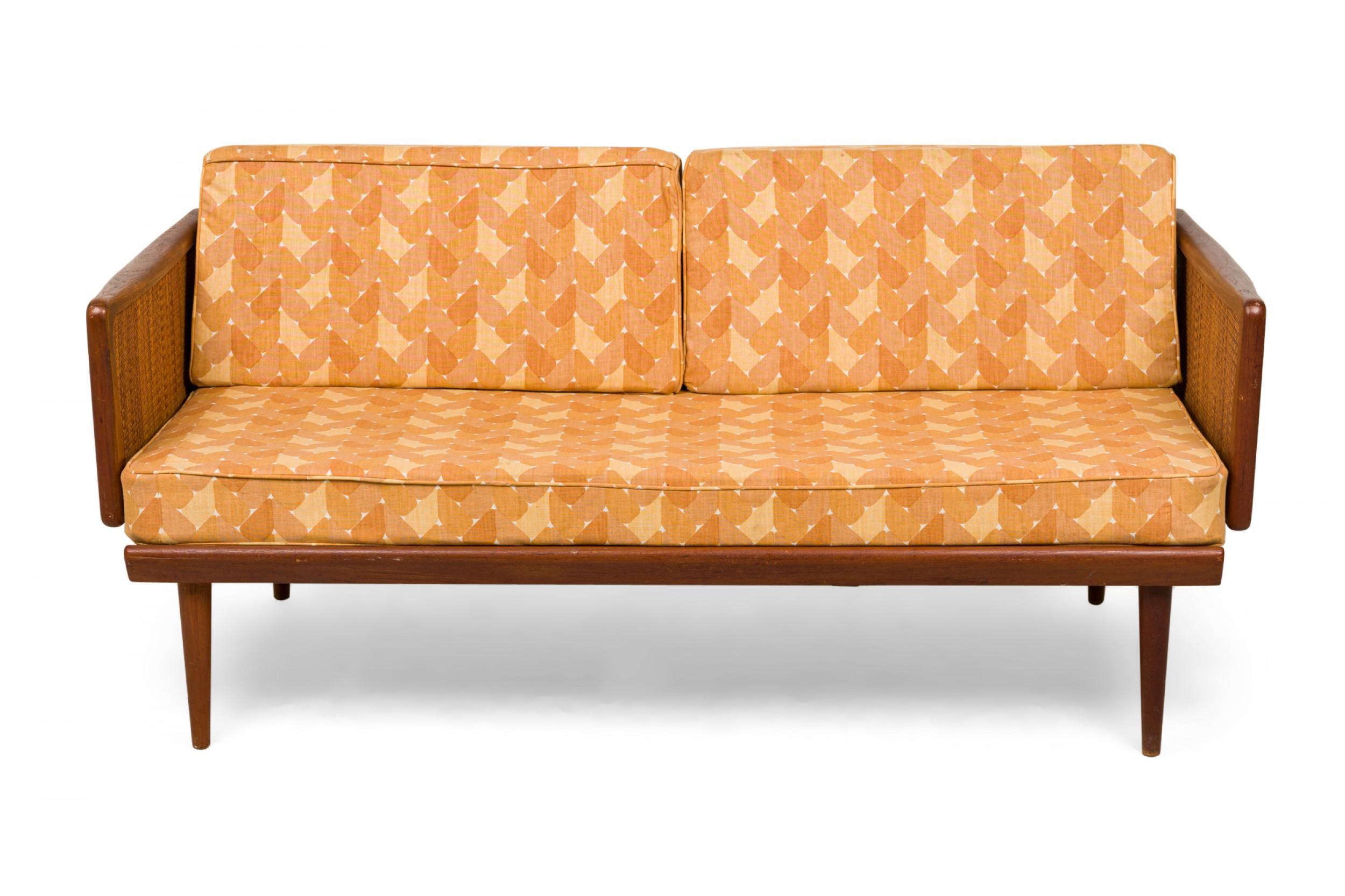 Danish Mid-Century expandable sleeper settee with a teak wood frame with caned sides, a solid seat cushion and two wedge-shaped removable back cushions in light and dark gold patterned upholstery, with hinged arms that fold down to create and