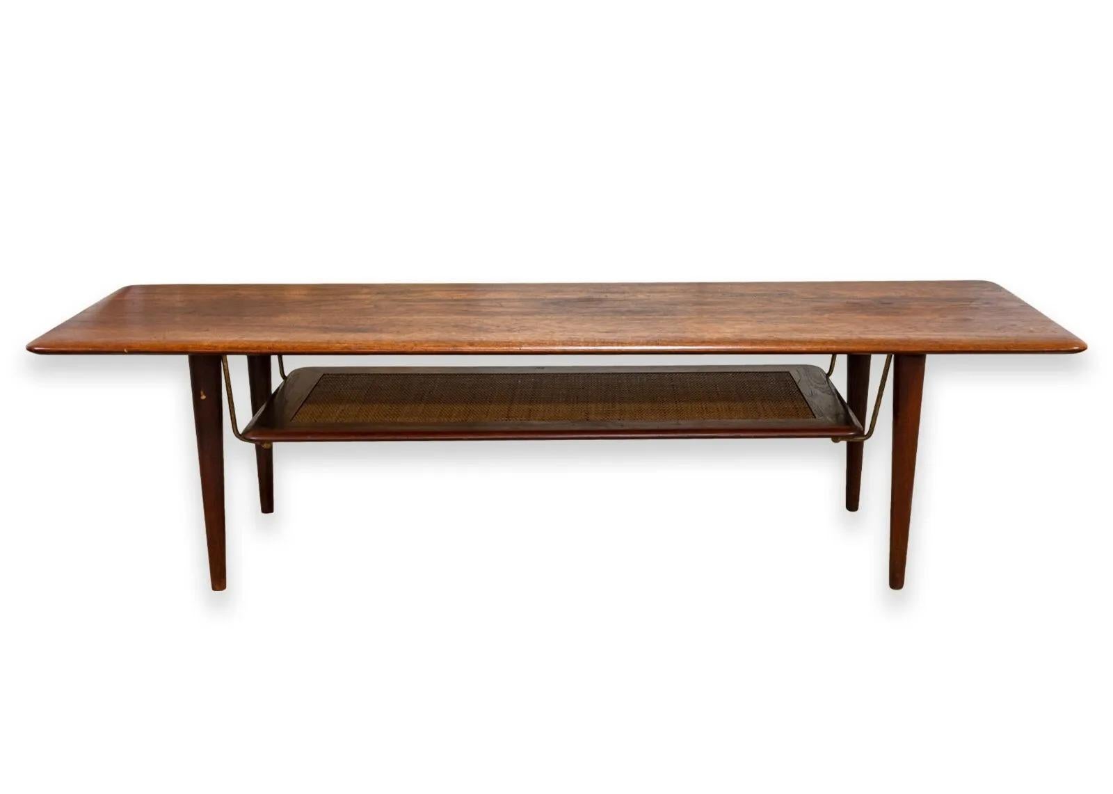 A Peter Hvidt for John Stuart teak coffee table. A stunning mid century modern coffee table constructed from a beautiful teak wood and accented with a cane weaving. This table features a two tier design. The lower tier hangs down via brass