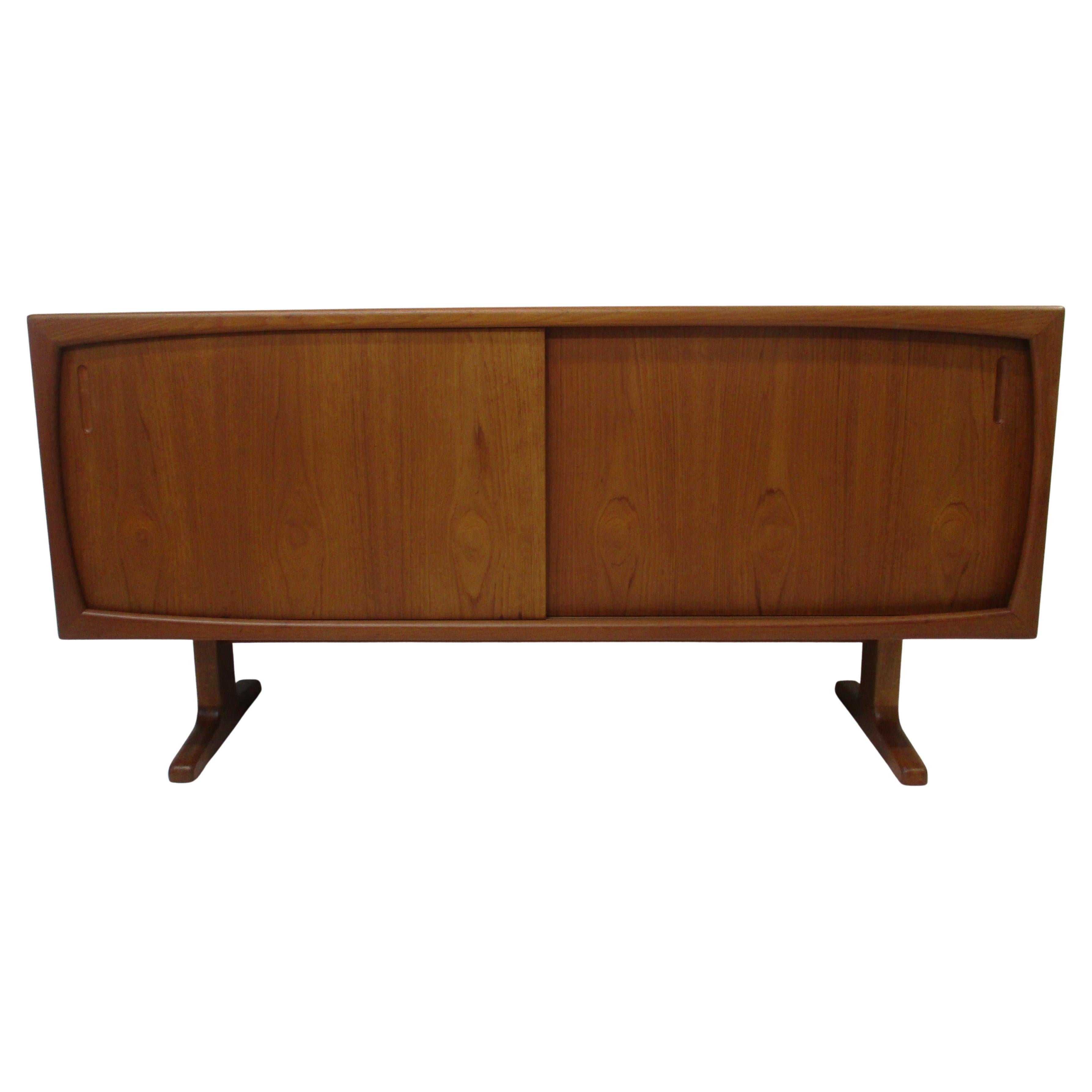Una credenza in teak dal design unico e affusolato con due ante scorrevoli con tiretti incorporati che contengono due cassetti superiori più piccoli e un unico ripiano regolabile. Poggiato su gambe scultoree e dotato di splendide ante in teak