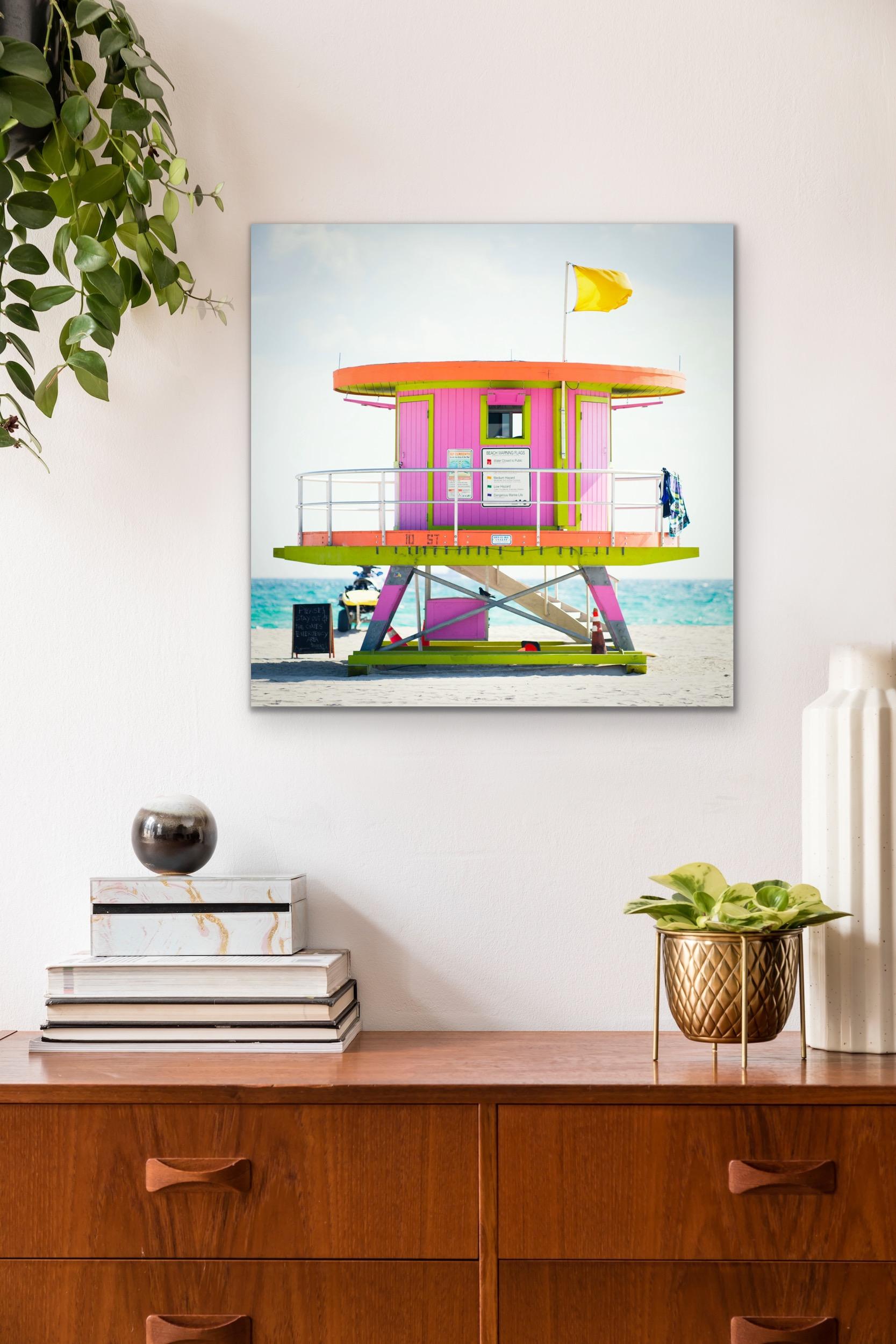 This contemporary coastal photograph by Peter Mendelson features a bright pink, orange, and lime green lifeguard stand on Miami Beach in Florida. The modern-looking, circular structure, sits on the sand with a bright blue and lightly cloudy sky