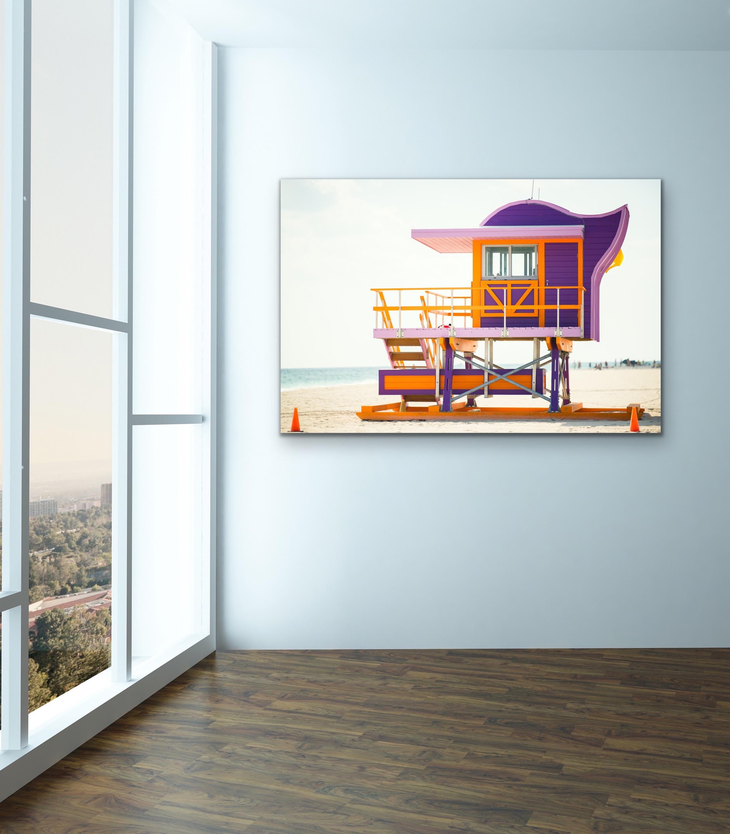 This contemporary coastal photograph by Peter Mendelson depicts a side view of a bright violet, pink, and orange Miami lifeguard stand on the beach. A light blue ocean can be seen behind the stand, with a light, neutral blue sky above it.