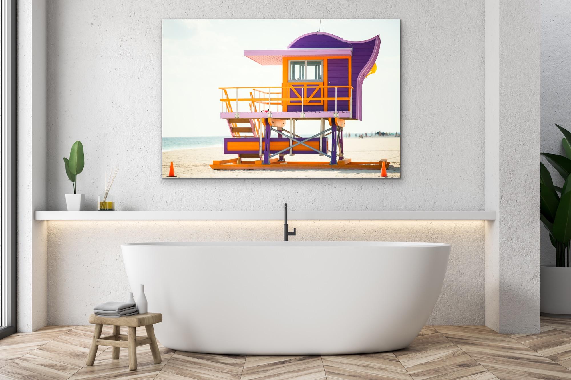 This contemporary coastal photograph by Peter Mendelson depicts a side view of a bright violet, pink, and orange Miami lifeguard stand on the beach. A light blue ocean can be seen behind the stand, with a light, neutral blue sky above it.