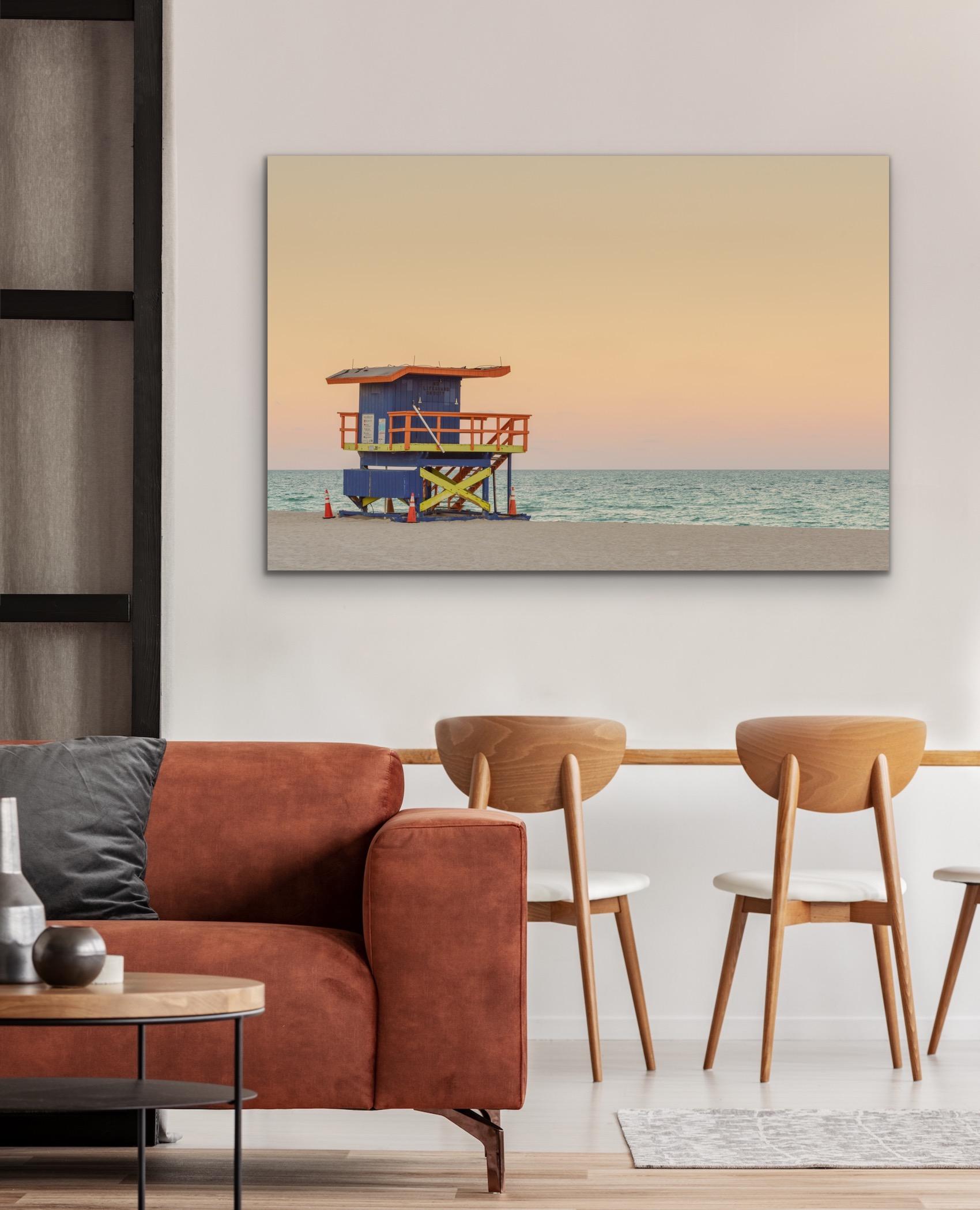 This contemporary coastal photograph by Peter Mendelson features a blue, orange, and yellow painted lifeguard stand in Miami, looking out over a muted teal ocean and a pink and yellow sunset. It is part of a series of photographs taken of the famous