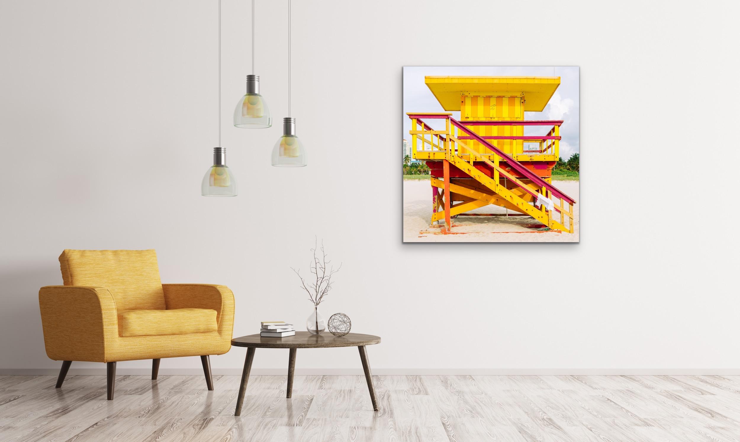 This contemporary coastal photograph by Peter Mendelson depicts a bright yellow, orange, and red lifeguard stand on Miami Beach in Florida. The structure features a yellow staircase with red railing that leads up to a landing where yellow and orange