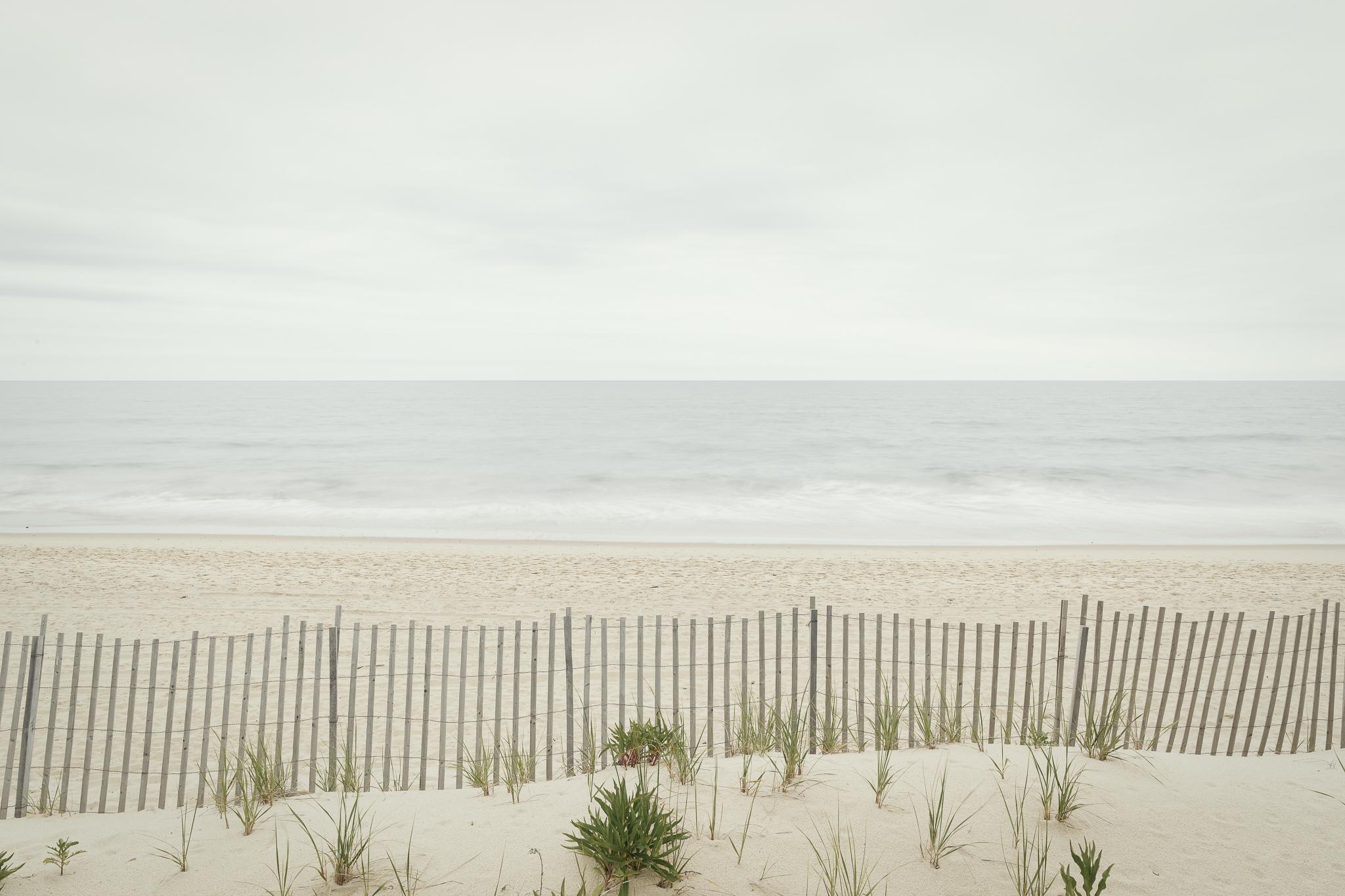 This contemporary coastal photograph features a light, neutral palette. It captures a New England beach scene, with a wooden fence in the sand in the foreground, and the ocean shore behind it, with a soft and serene overall aesthetic. 

This is a